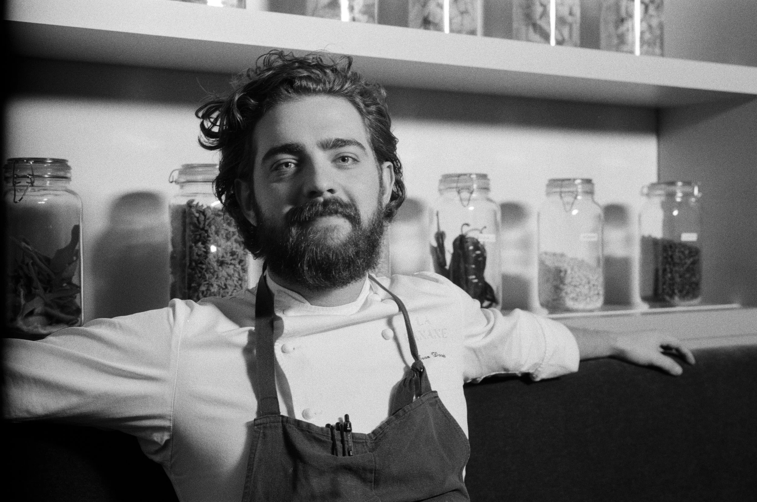 A male chef with a beard and curly hair leaning against a countertop, wearing a white chef's jacket and apron, with jars of ingredients on a shelf behind him.