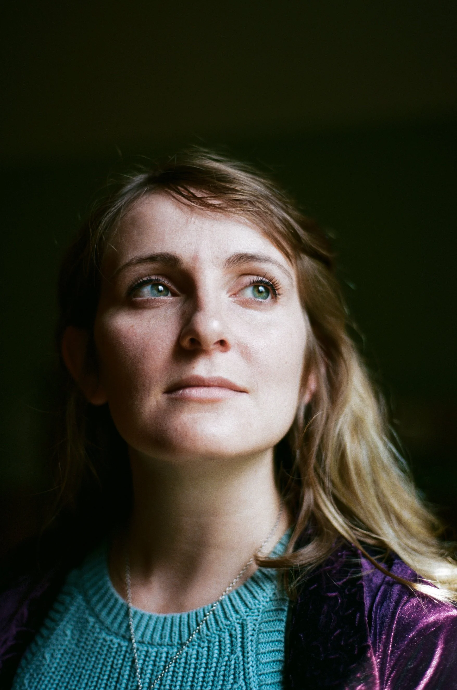 Close-up portrait of a woman with blue eyes, light brown hair, wearing a teal knitted sweater and a purple velvet jacket, looking thoughtfully to the side.