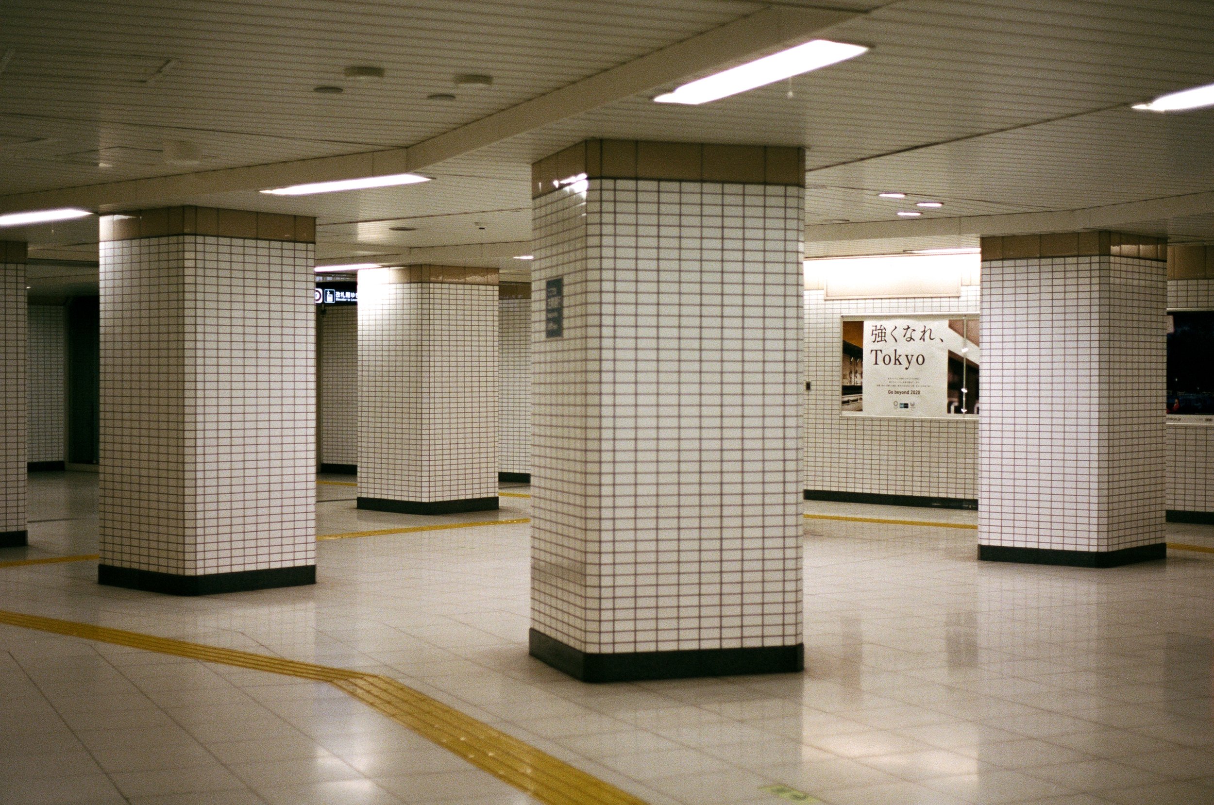 An underground subway station with white tiled pillars and floors, illuminated by ceiling lights. A sign with Japanese and English text reads "Go beyond 2020," with "Tokyo" written beneath.