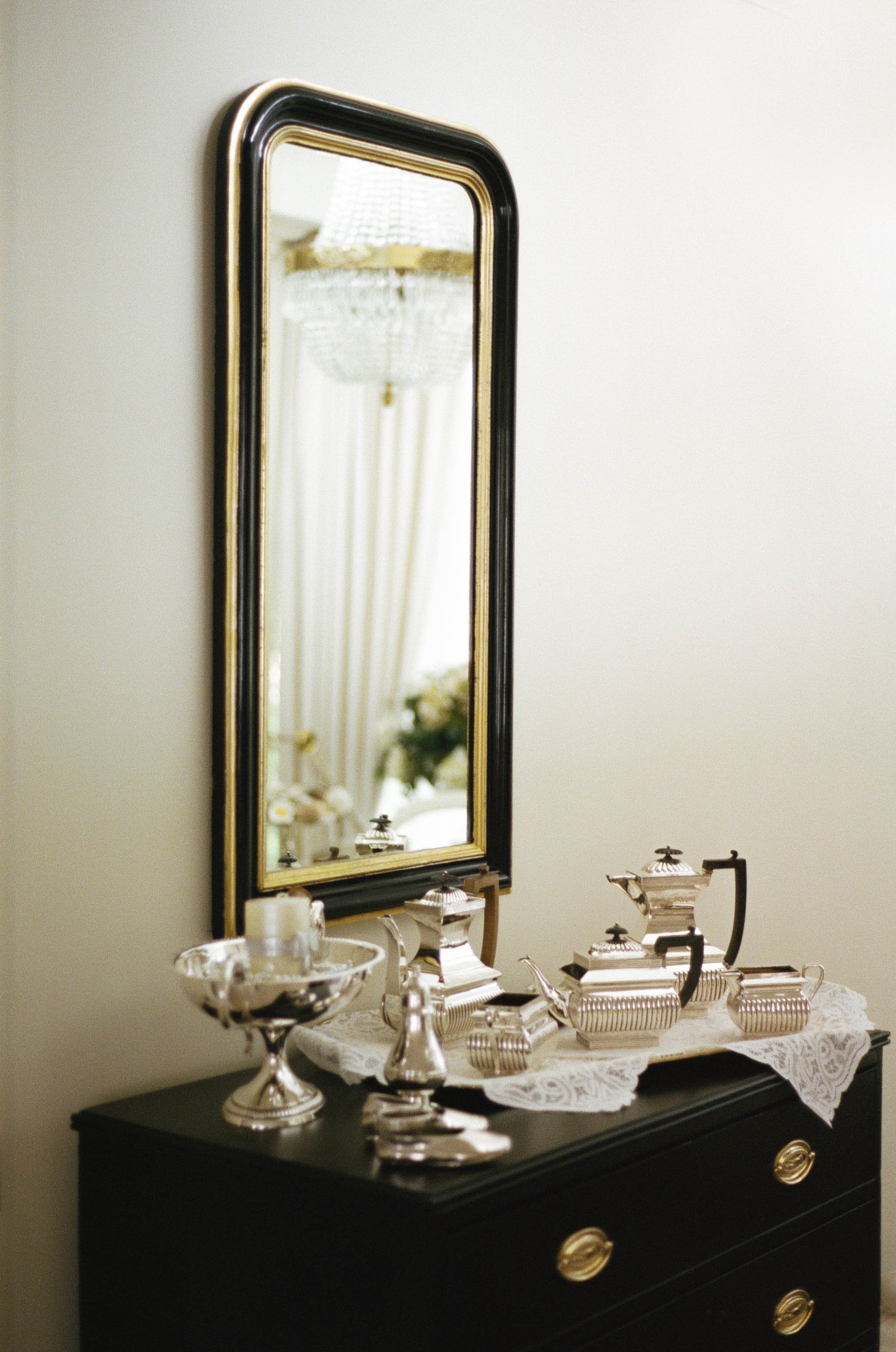 A black dresser with gold handles, decorated with silver tea sets and a lace cloth, a mirror above reflecting a chandelier and curtains.