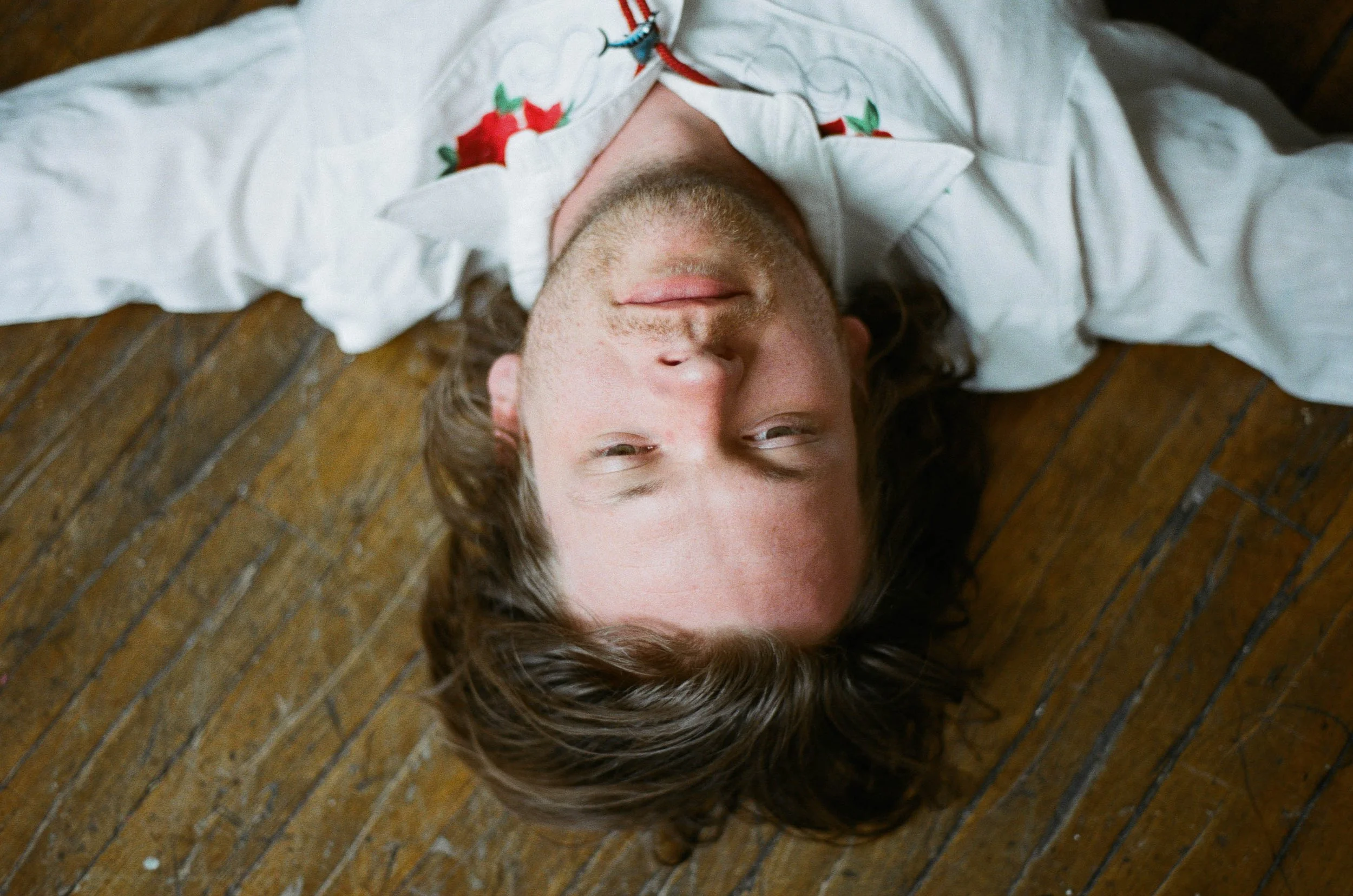 A man with medium-length brown hair and freckles lies on a wooden floor, head tilted back and eyes partially closed, wearing a white shirt with embroidered floral details.