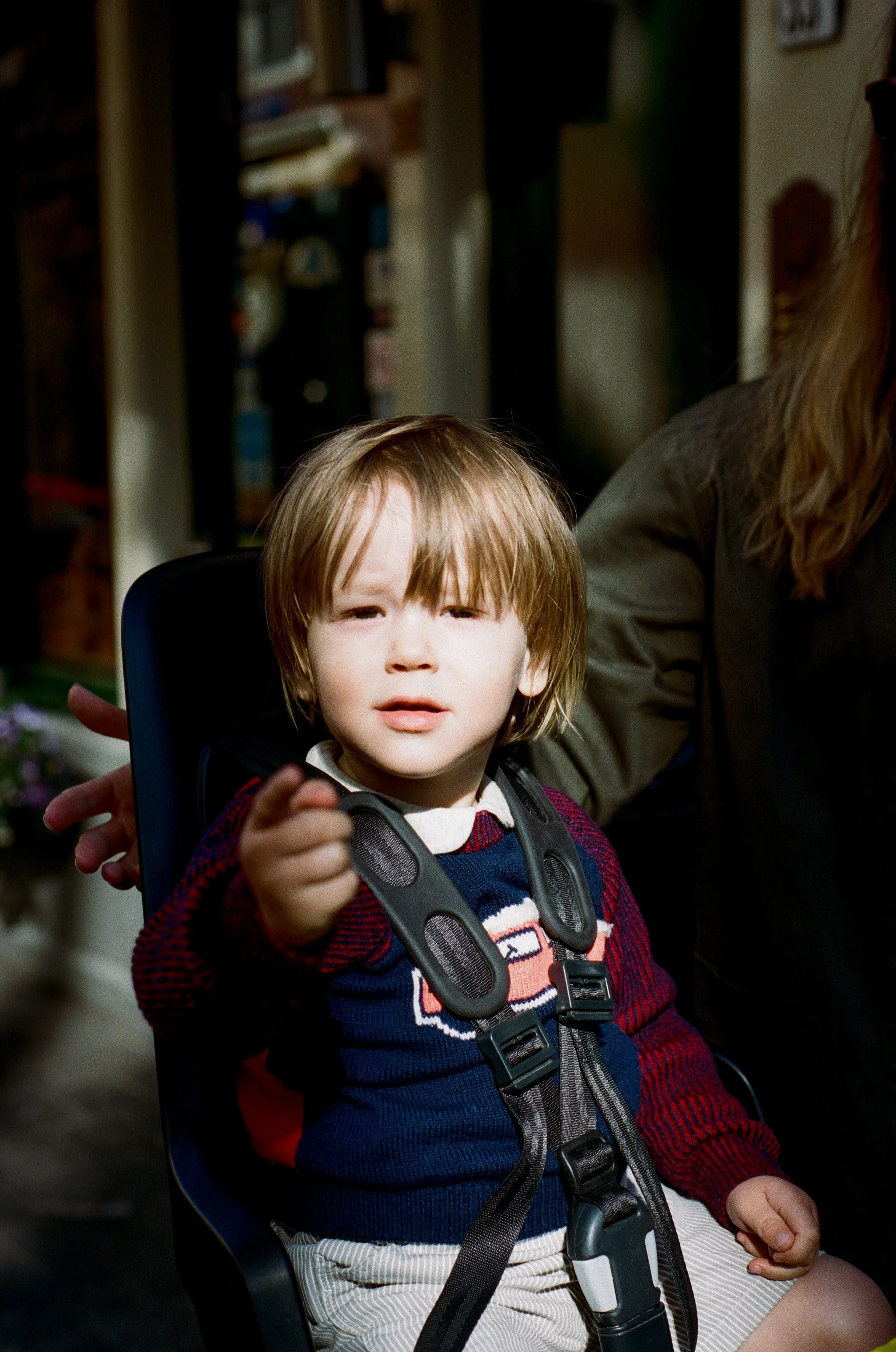 A young boy with light brown hair in a high chair, pointing at the camera with a serious expression, wearing a sweater with a car pattern and a harness.