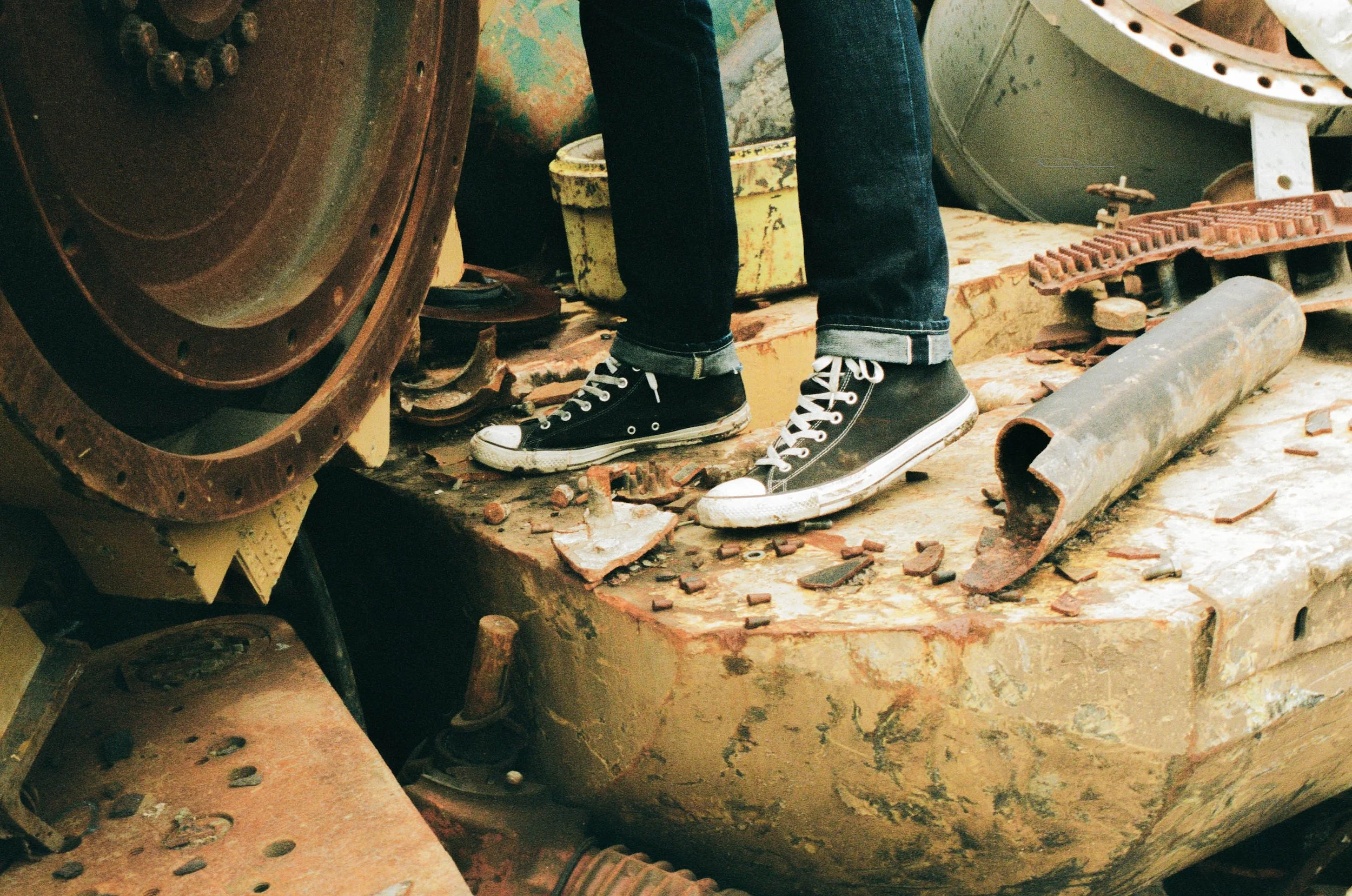 Person wearing black Converse sneakers standing on rusted, damaged metal surface surrounded by debris and machinery parts.