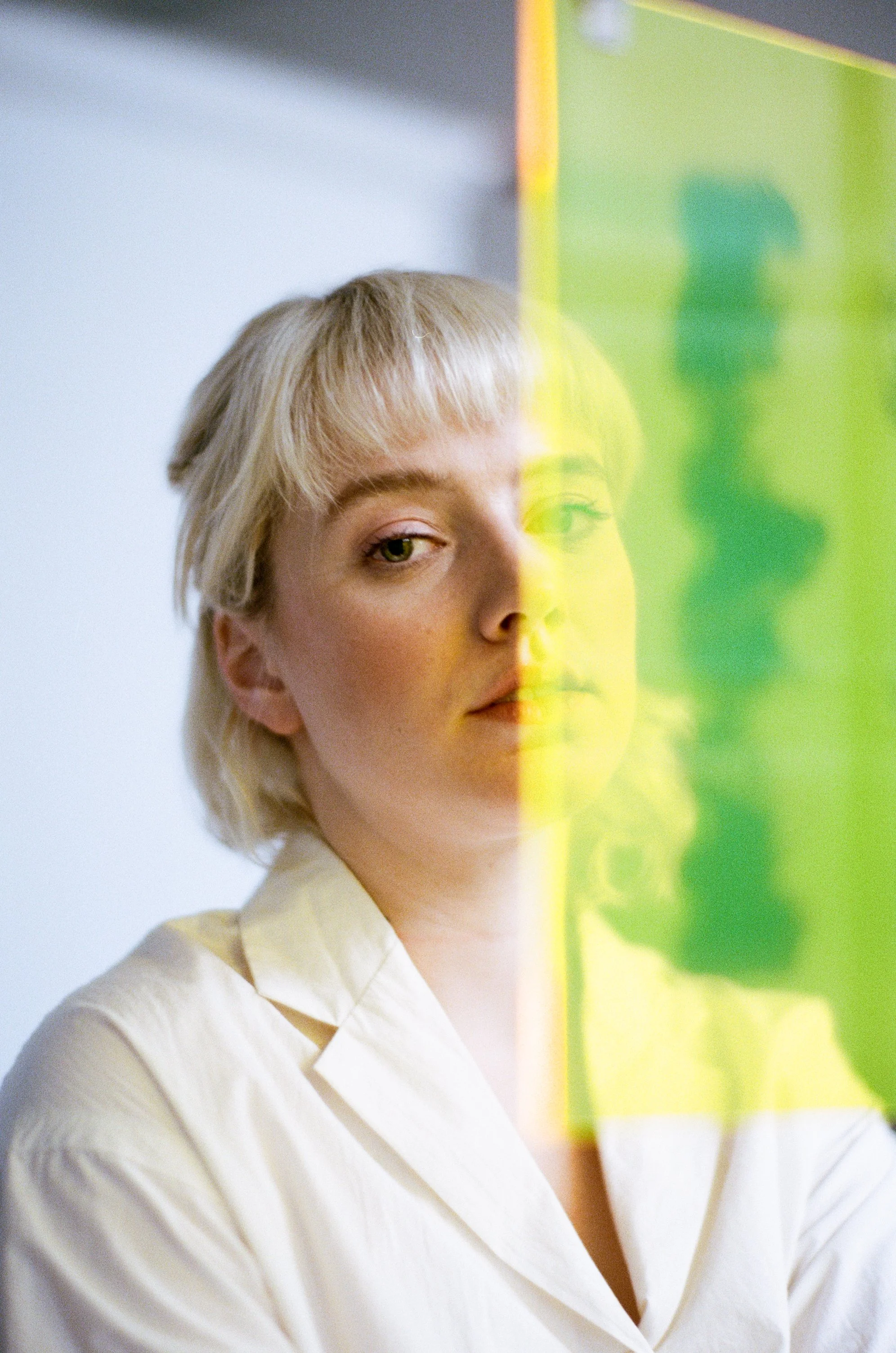 A young woman with short blonde hair and light makeup looks at a neon yellow-green sign or glass panel that partially obscures her face.