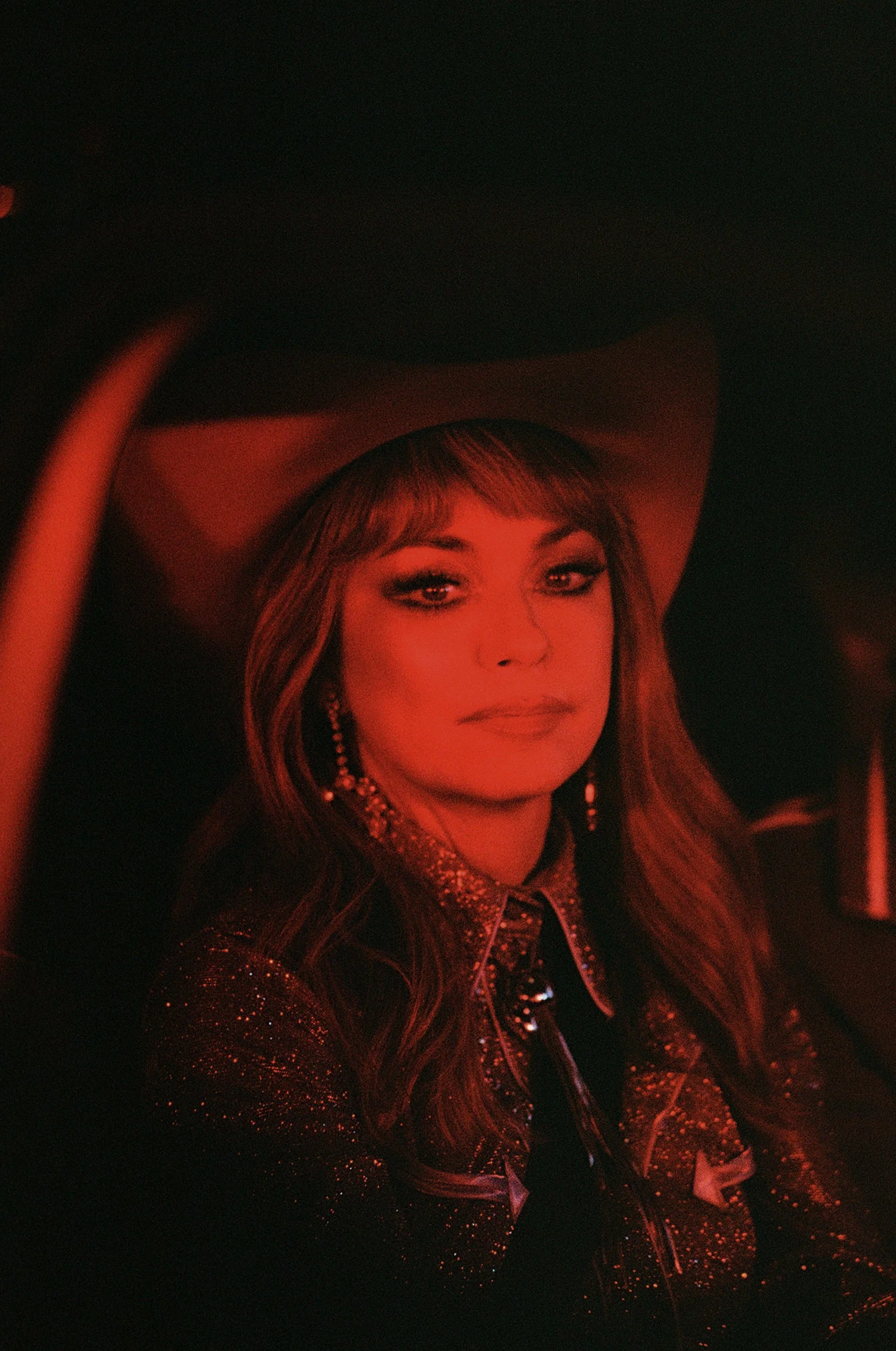 A woman with styled hair and makeup, wearing a cowboy hat and a sparkly jacket, sitting in a dimly lit setting with a red hue.