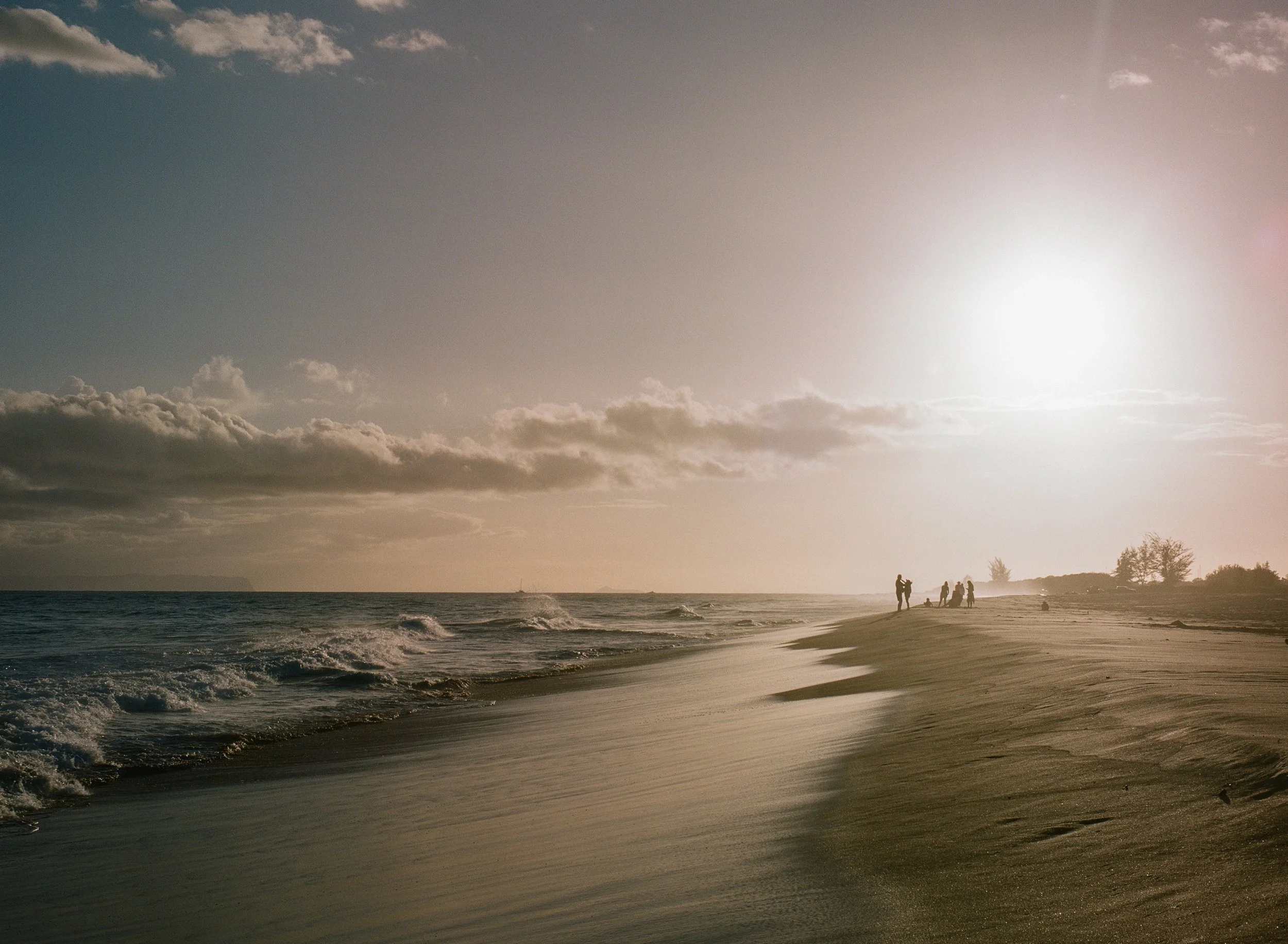 Beach at sunset with a few people walking along the shoreline and trees in the distance.