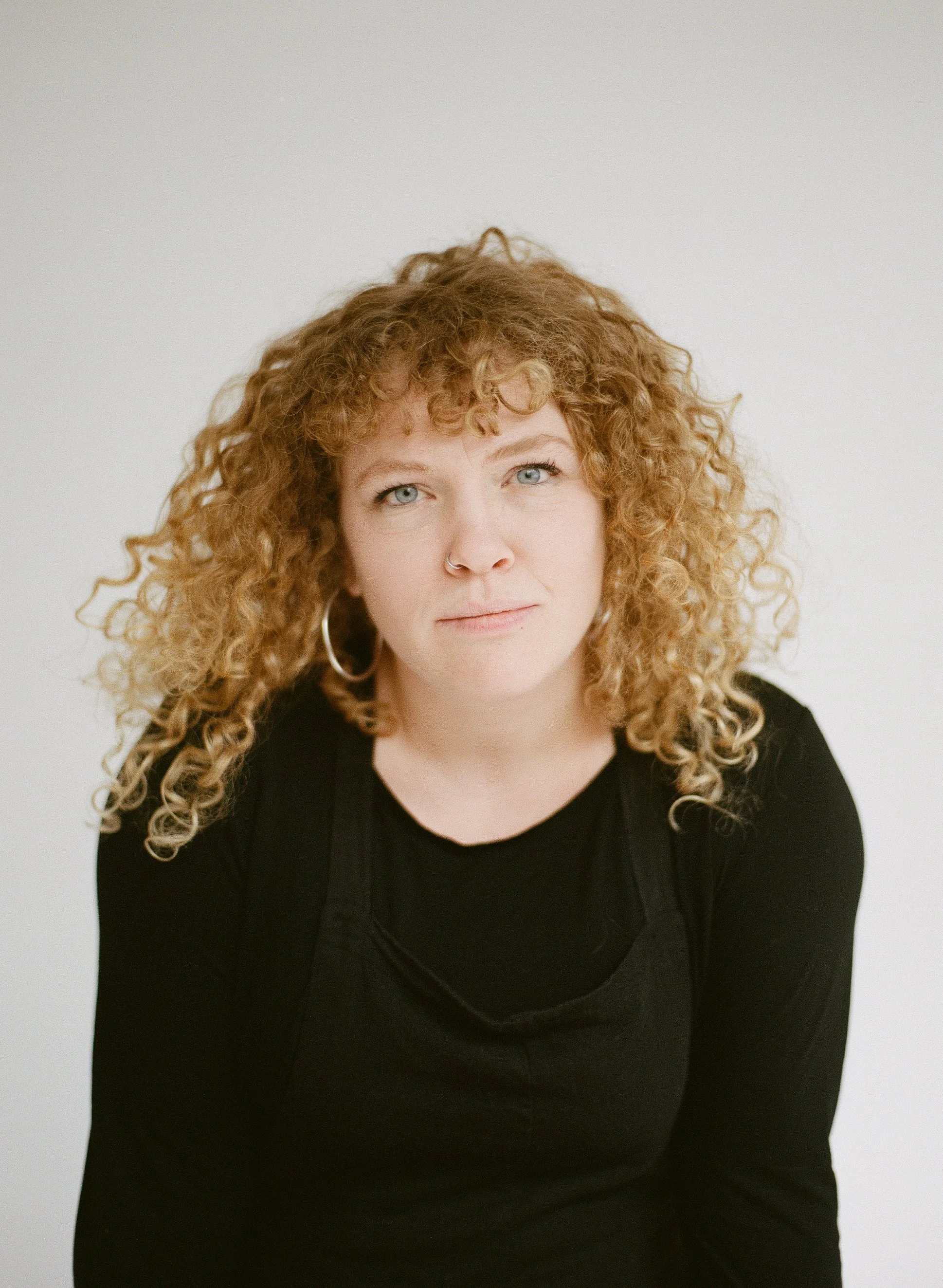 A woman with curly red hair, blue eyes, wearing hoop earrings, a nose ring, and a black top, looking directly at the camera against a plain white background.