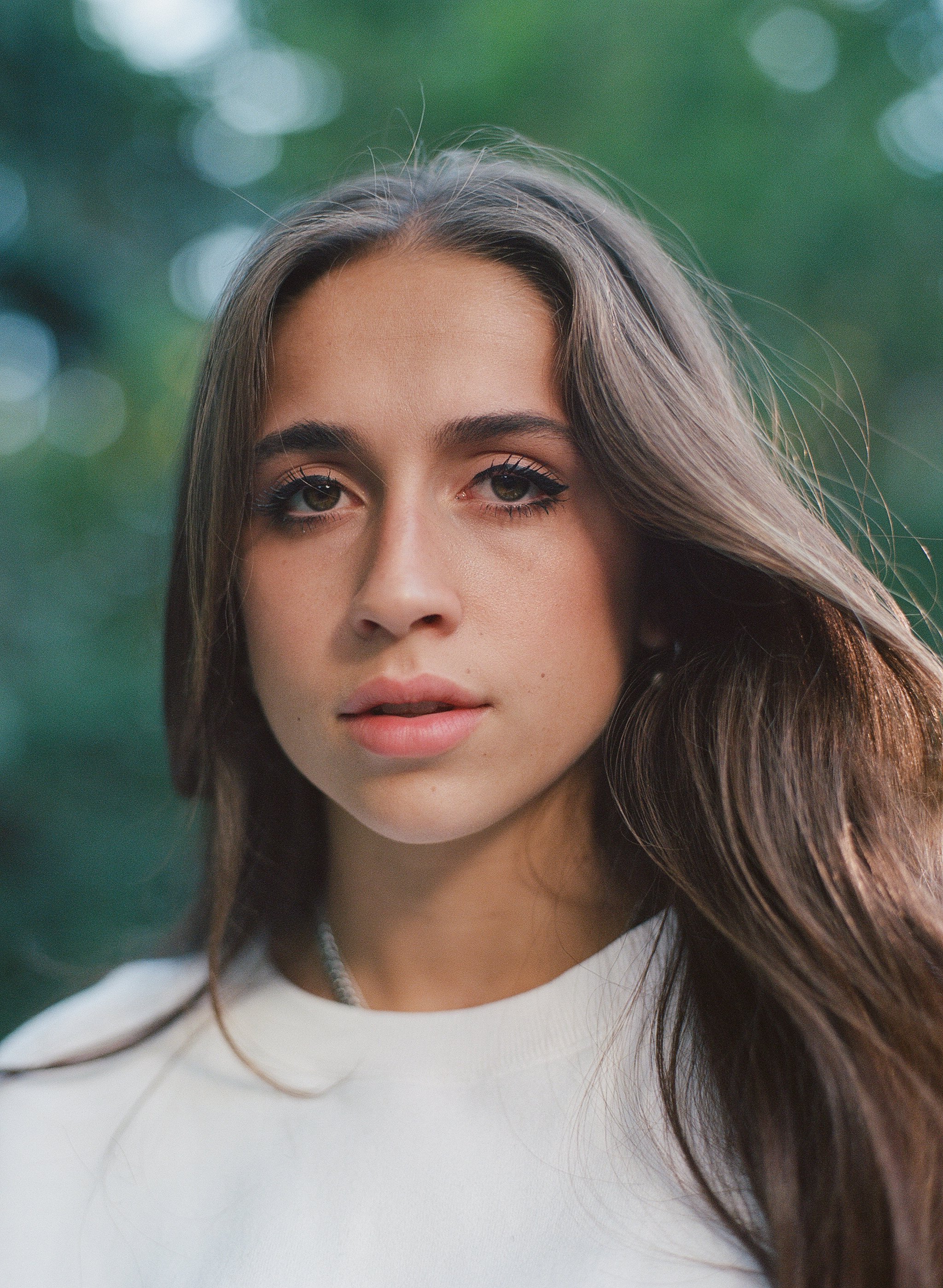 Close-up portrait of a young woman with long brown hair, wearing a white top, outdoors with blurred green background.