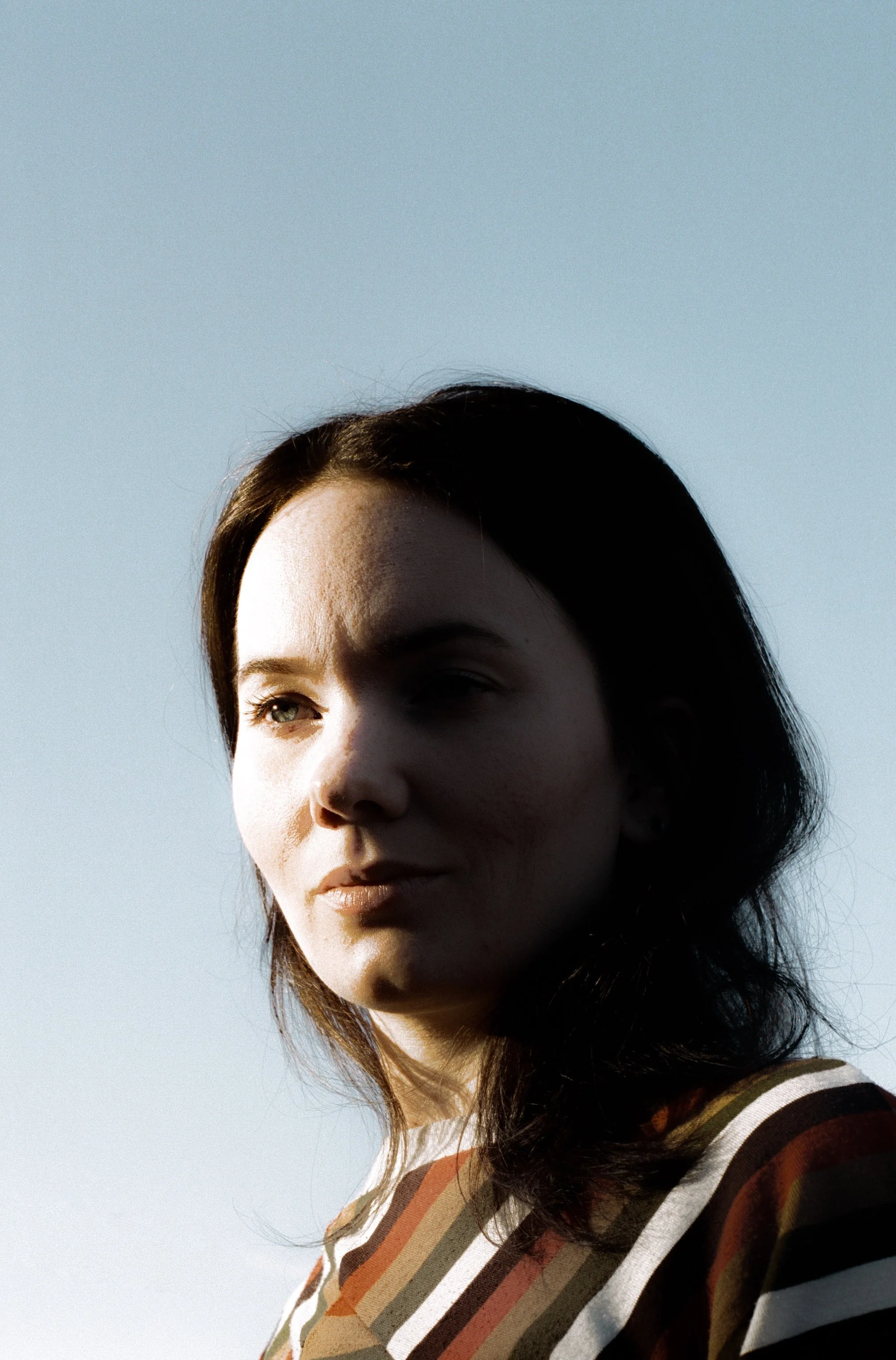 A woman with dark hair and light skin, wearing a striped shirt, standing outdoors against a clear blue sky with sunlight partially illuminating her face.