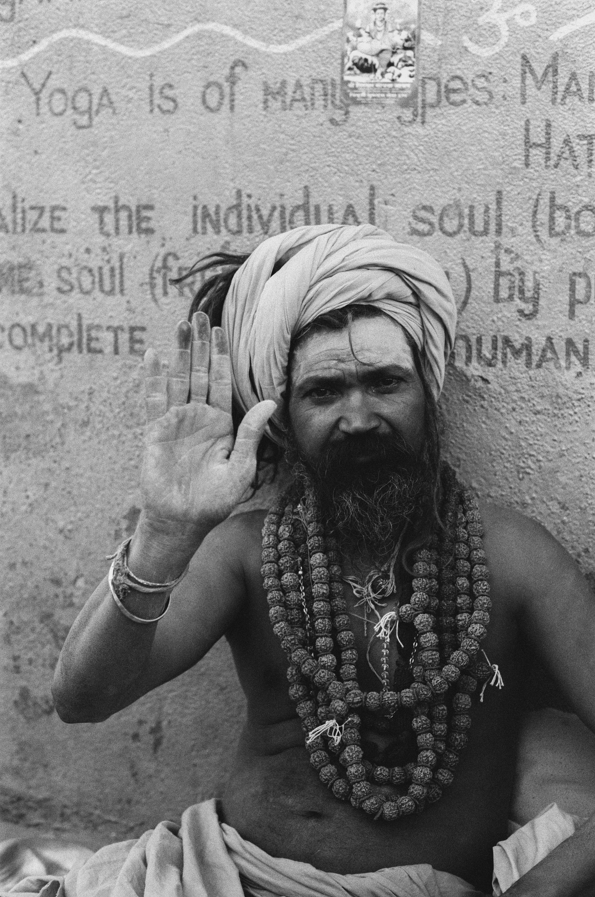 A bearded man wearing multiple necklaces and a turban, raising his right hand in a gesture of blessing or greeting, standing against a textured wall with partially visible text and an image at the top.