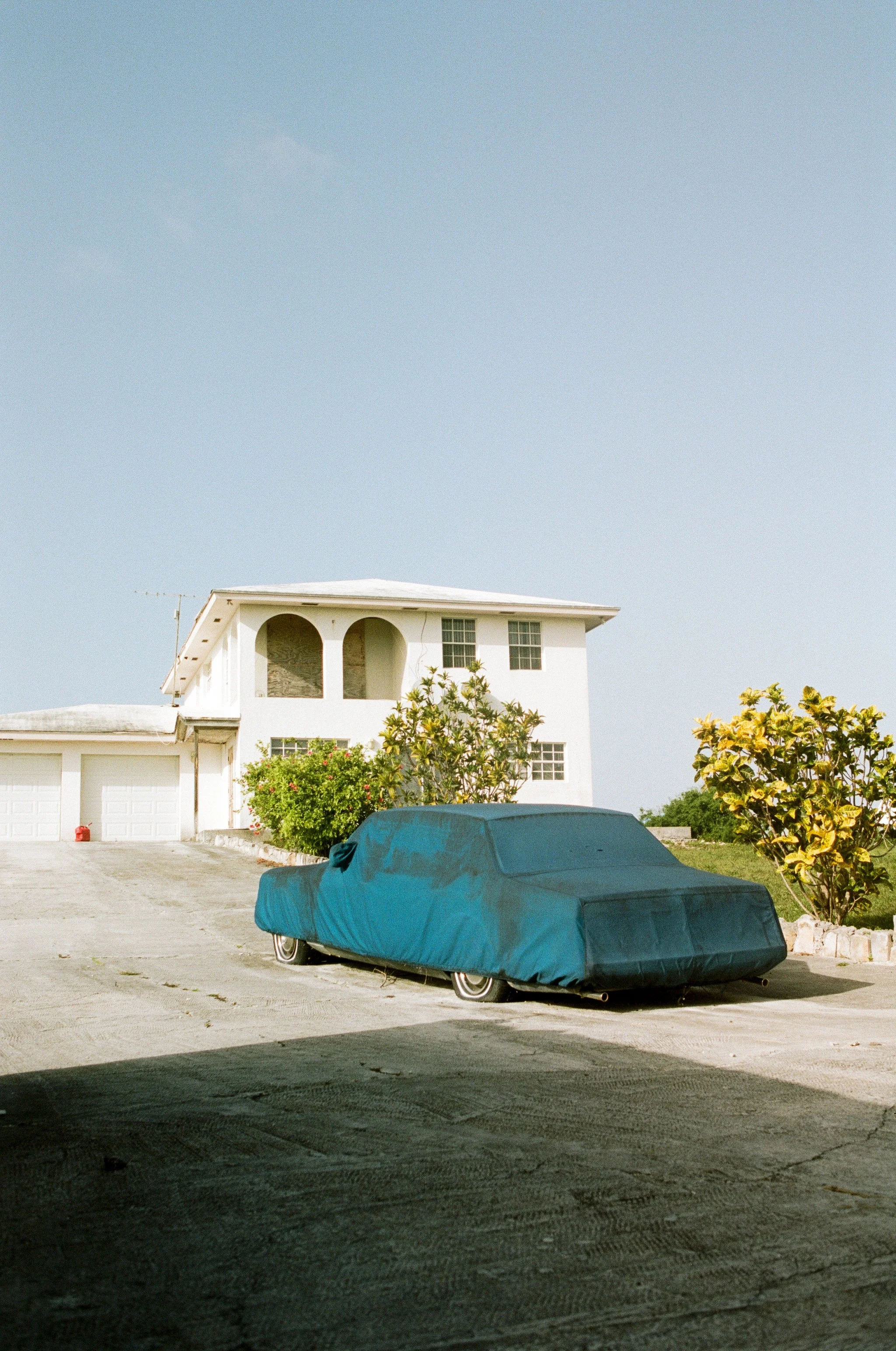A white two-story house with a driveway and a car covered with a blue car cover in front. There are bushes and trees in the yard, and the sky is clear.