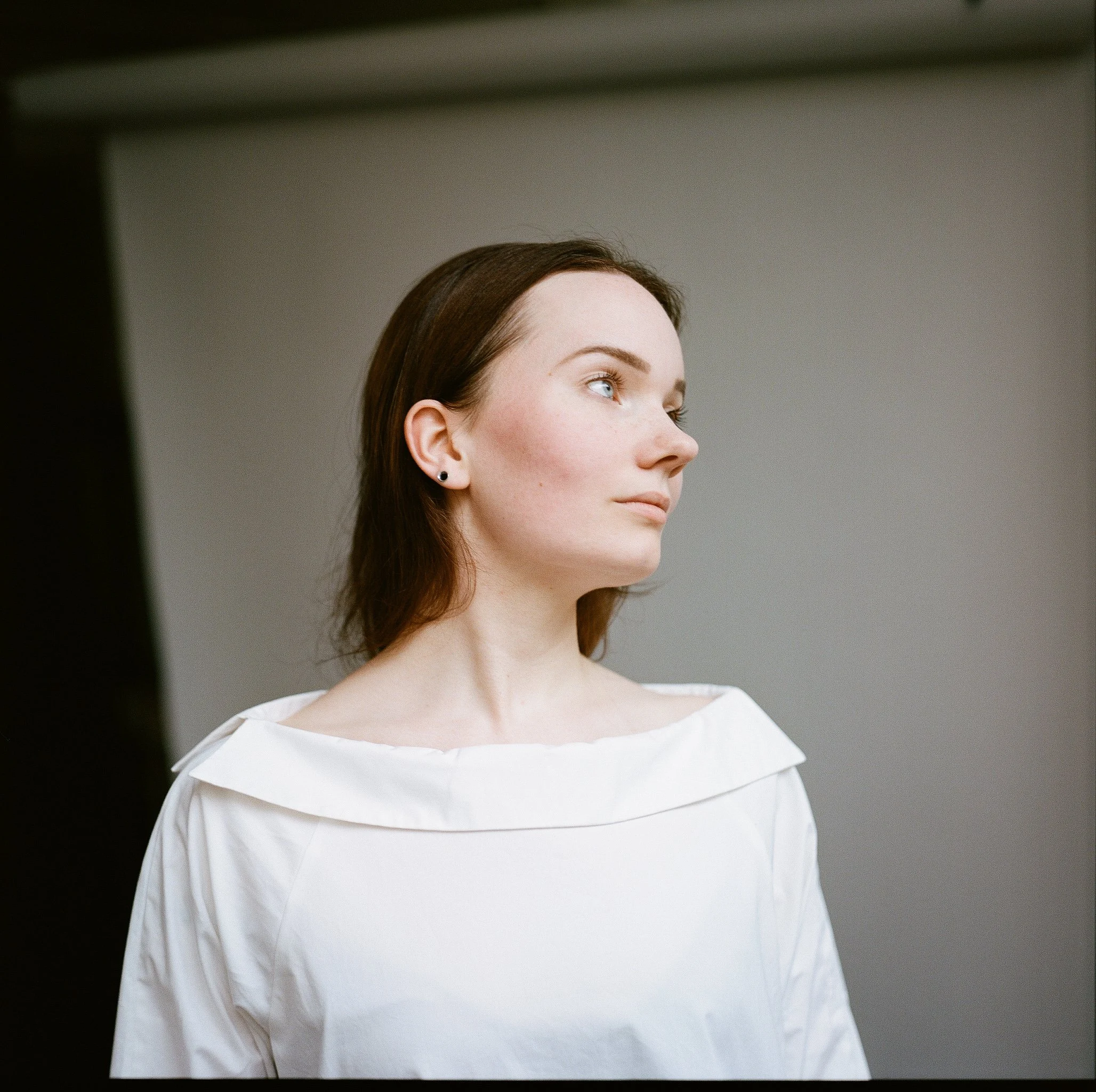A young woman with long brown hair and blue eyes looking to the right, wearing a white off-shoulder top, standing against a plain background.