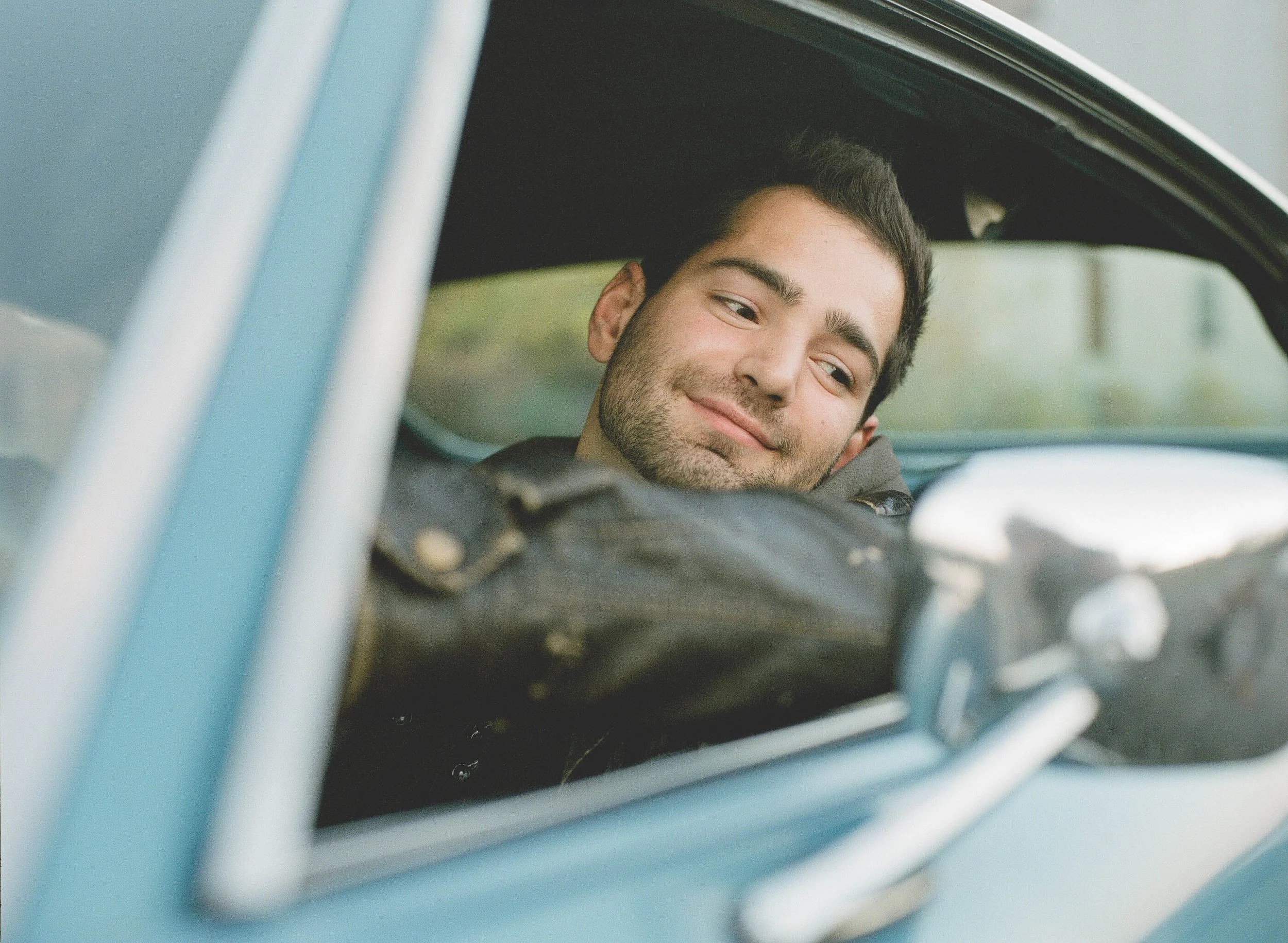 Young man with dark hair and beard relaxing inside a vintage car, looking out through the window with a smile.