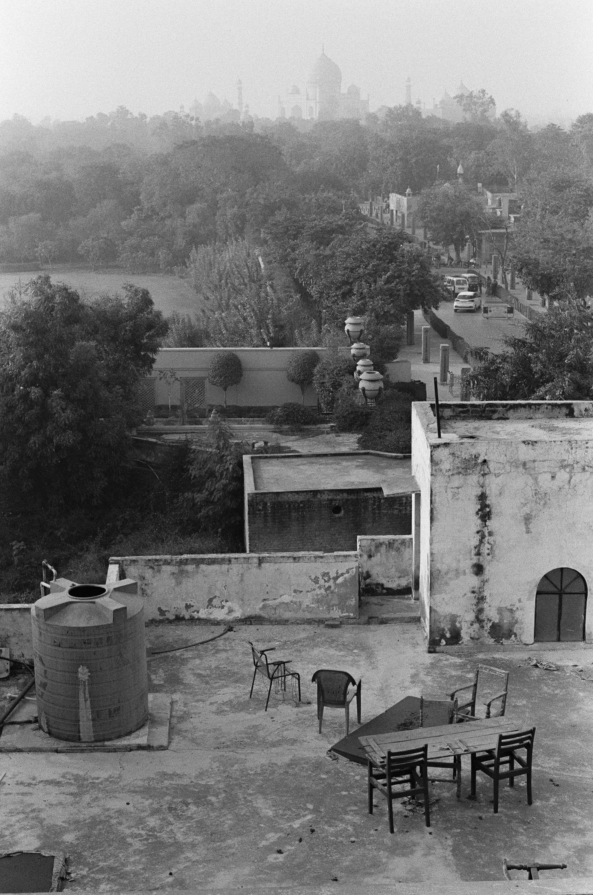 Black and white photo of a rooftop with chairs, a table, and a water tank, overlooking a cityscape with trees and a road.