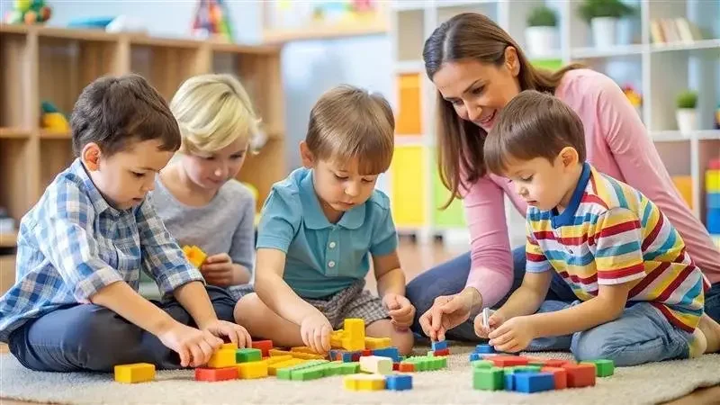 A woman and four young children sitting on the floor playing a colorful board game together in a classroom or playroom.