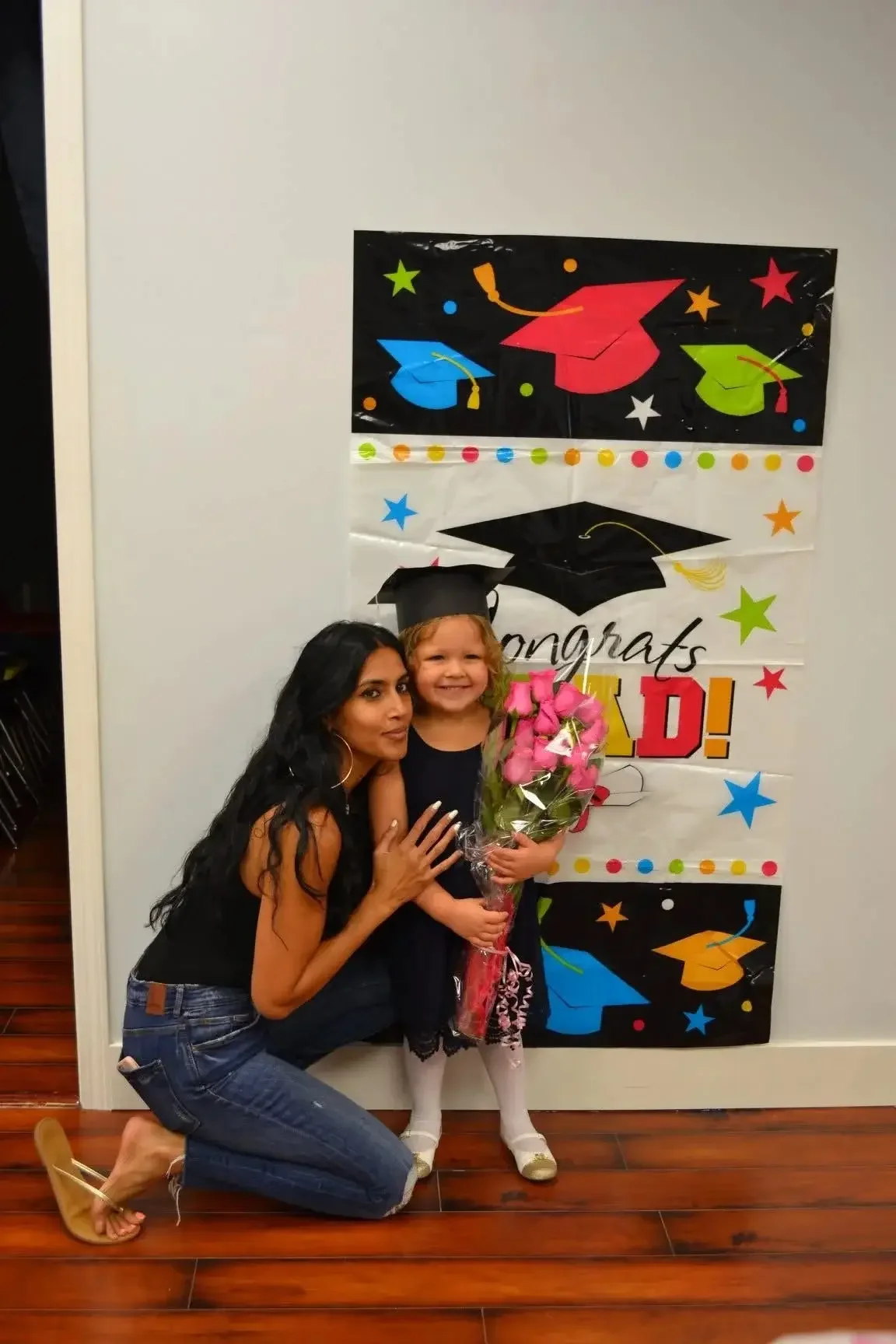A woman kneeling next to a young girl wearing a graduation cap, holding a bouquet of pink flowers, in front of an academic celebratory poster with colorful hats and stars.