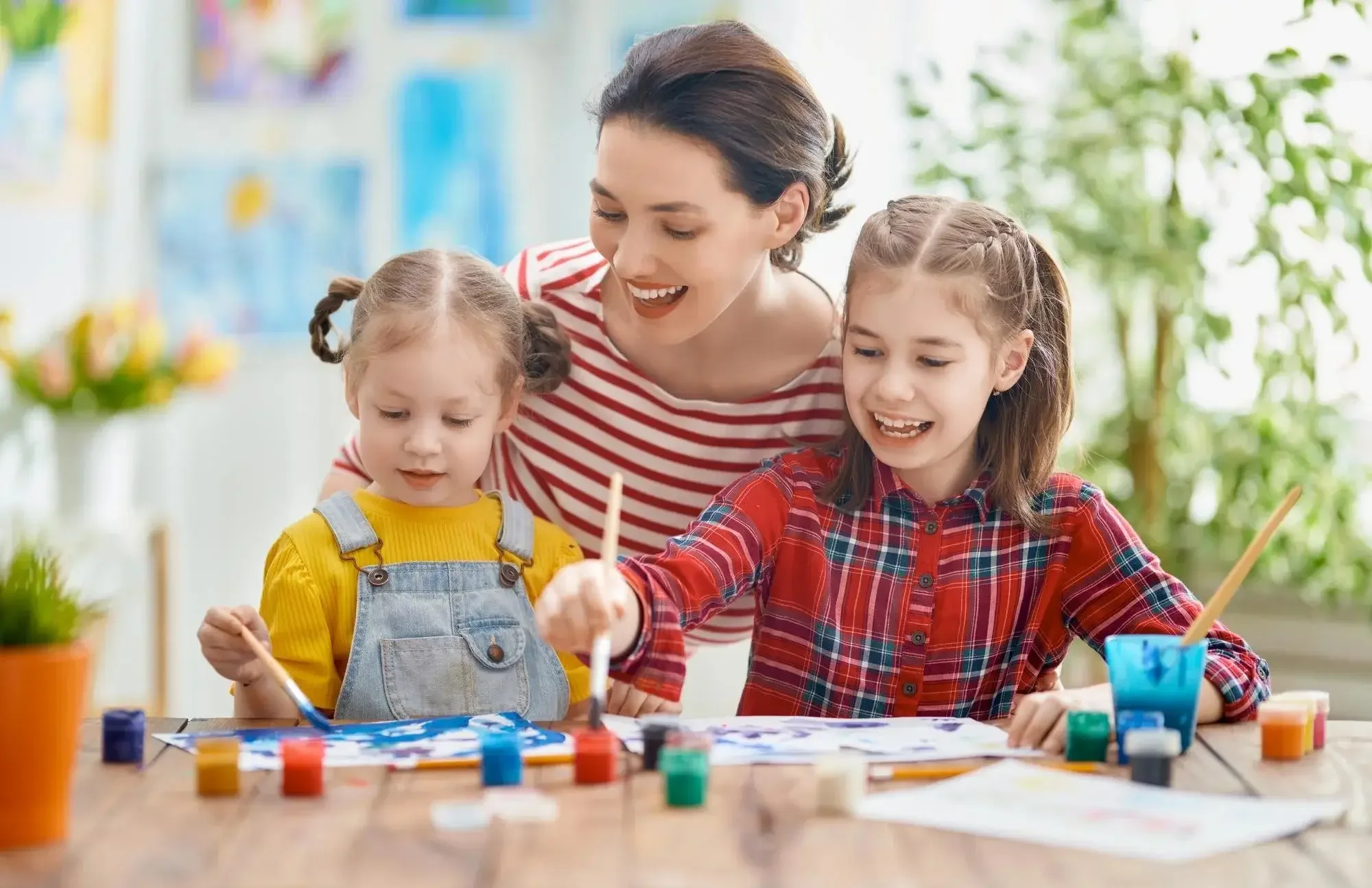 A woman and two young girls painting together at a table with paints and brushes, enjoying a creative activity indoors.