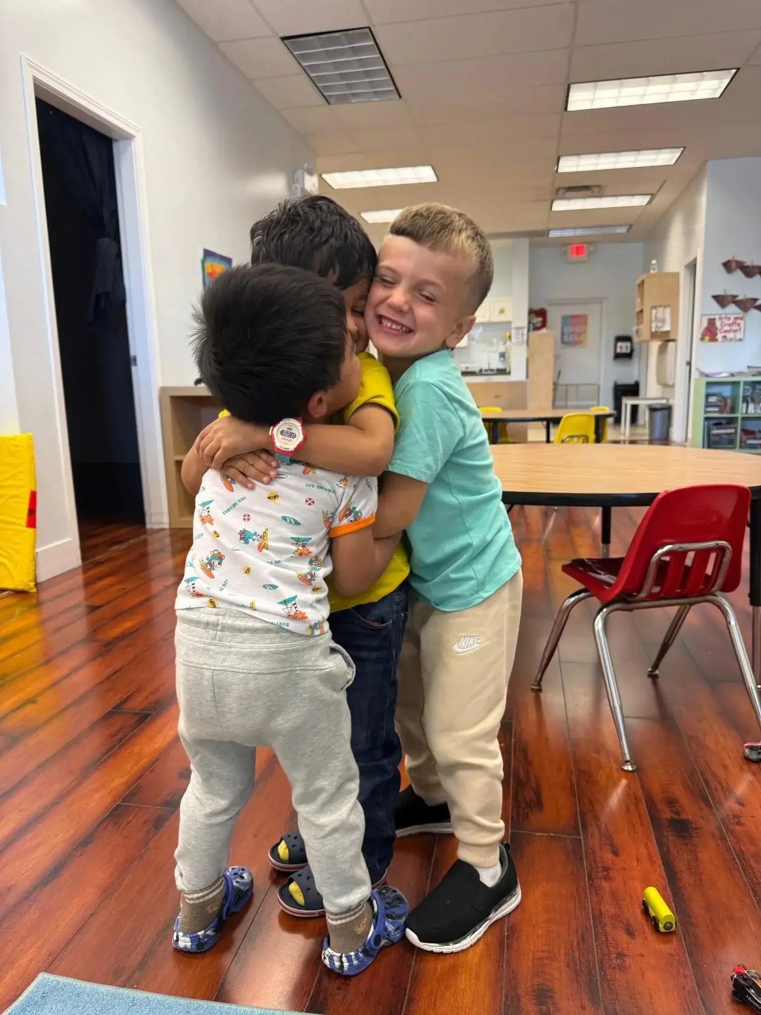 Three children sharing a group hug in a classroom with wooden flooring and tables.