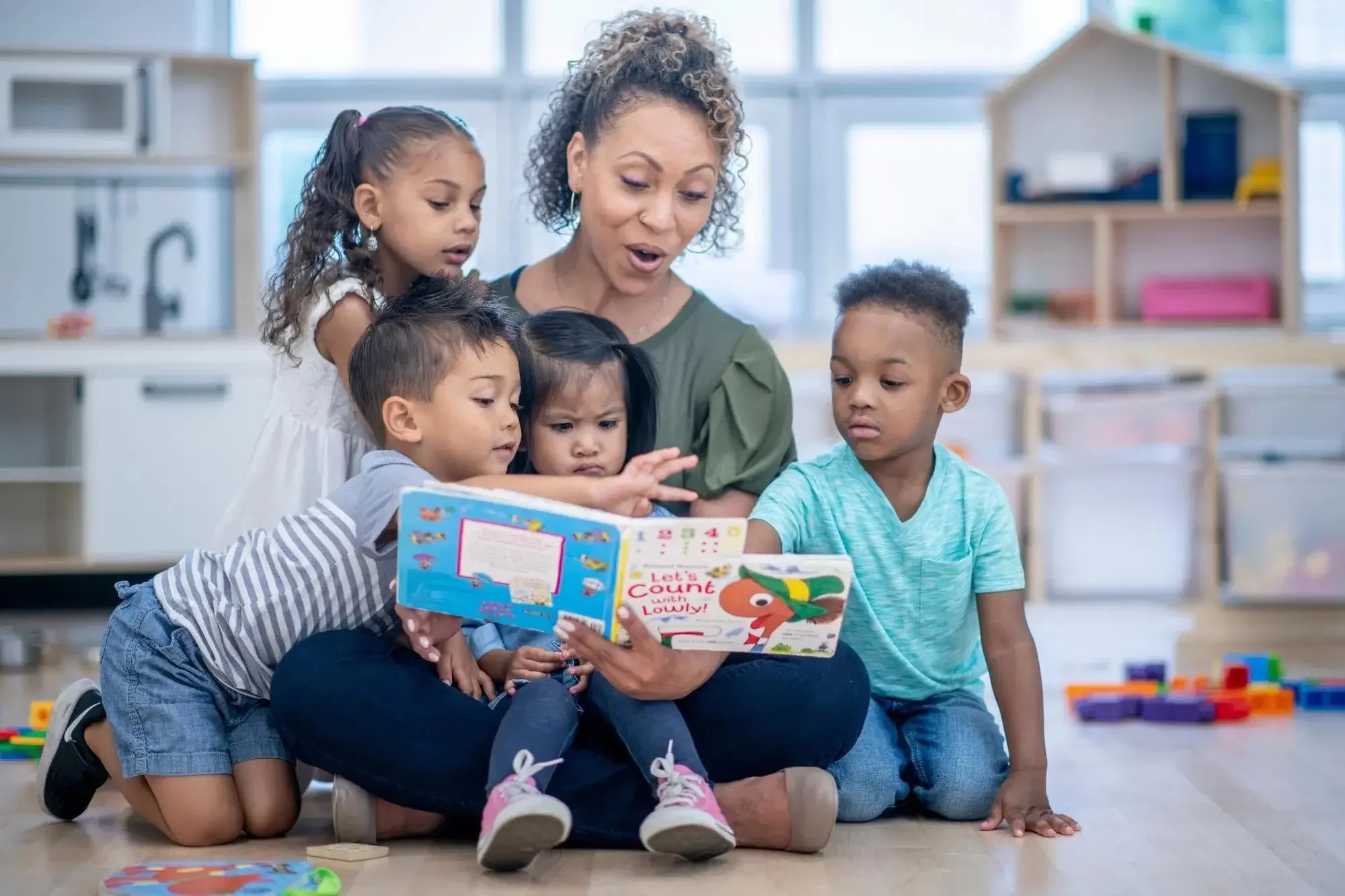 A teacher reading a children's book to a group of five diverse young children in a classroom.