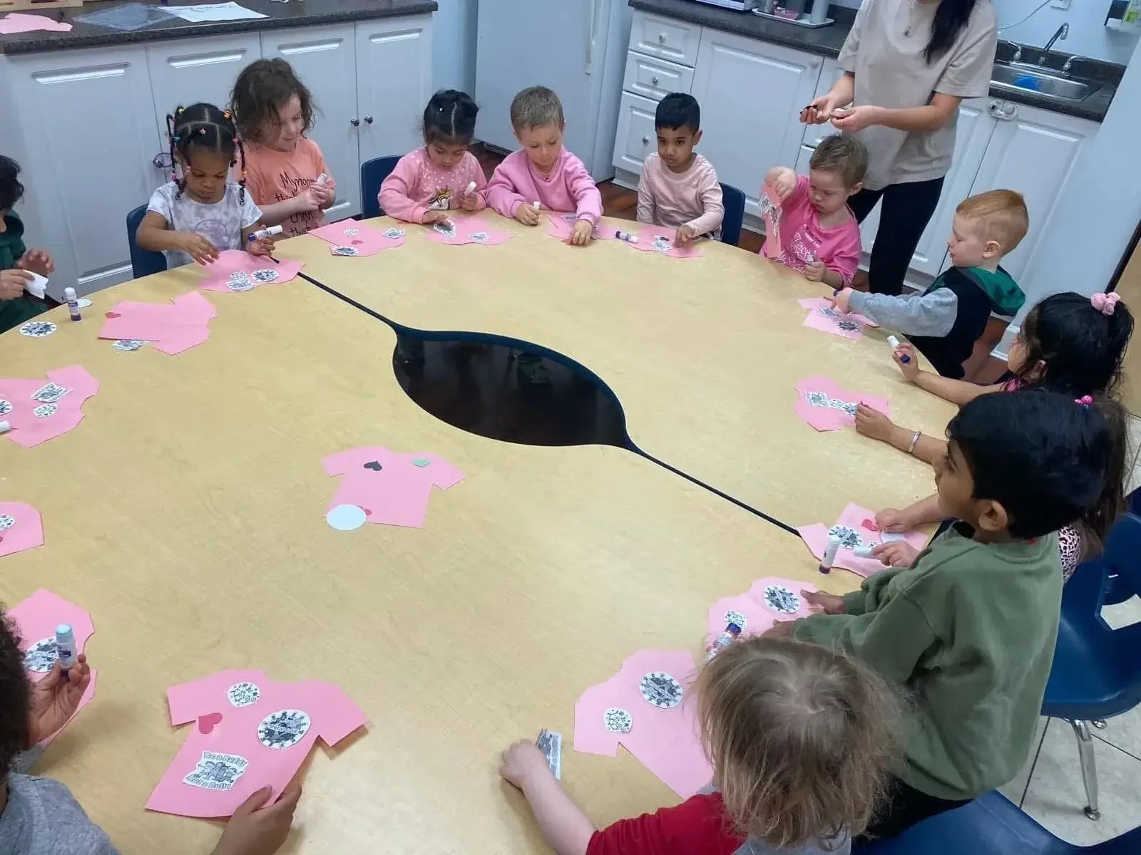 Children sitting around a large table with pink paper shirts, gluing and decorating while a teacher helps in a classroom.