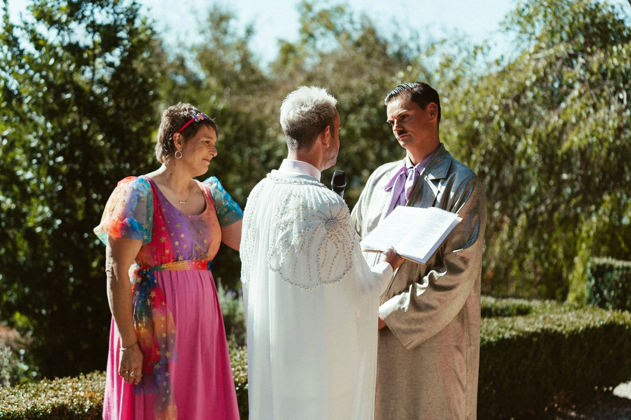 Dee Wild Celebrant, wearing a pink and rainbow dress is standing next to Keiran and Bryce as they recite their vows.