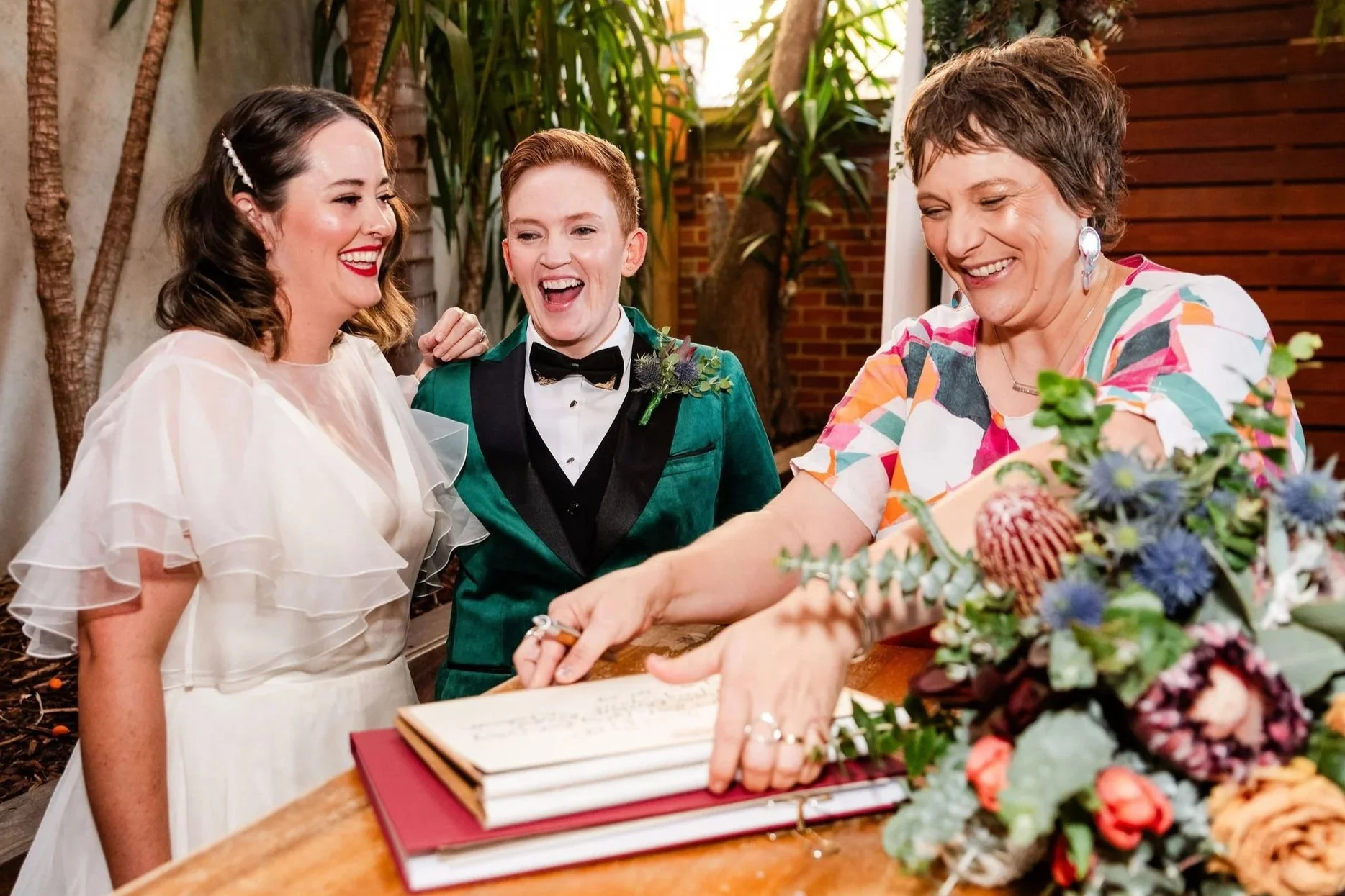 Dee Wild Celebrant is pictured with Tracy and Jell. Tracy is wearing a white wedding dress, Jell is wearing a green velvet suit. Dee Wild is wearing an orange and white dress. They are all laughing.
