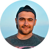 Portrait of a young man outdoors with a blue sky background, wearing a backwards baseball cap and a dark t-shirt.