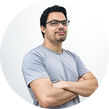 A man with glasses and a beard, wearing a light gray T-shirt, standing with arms crossed against a plain white background.