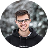 A young man with glasses smiling outdoors in a winter setting.