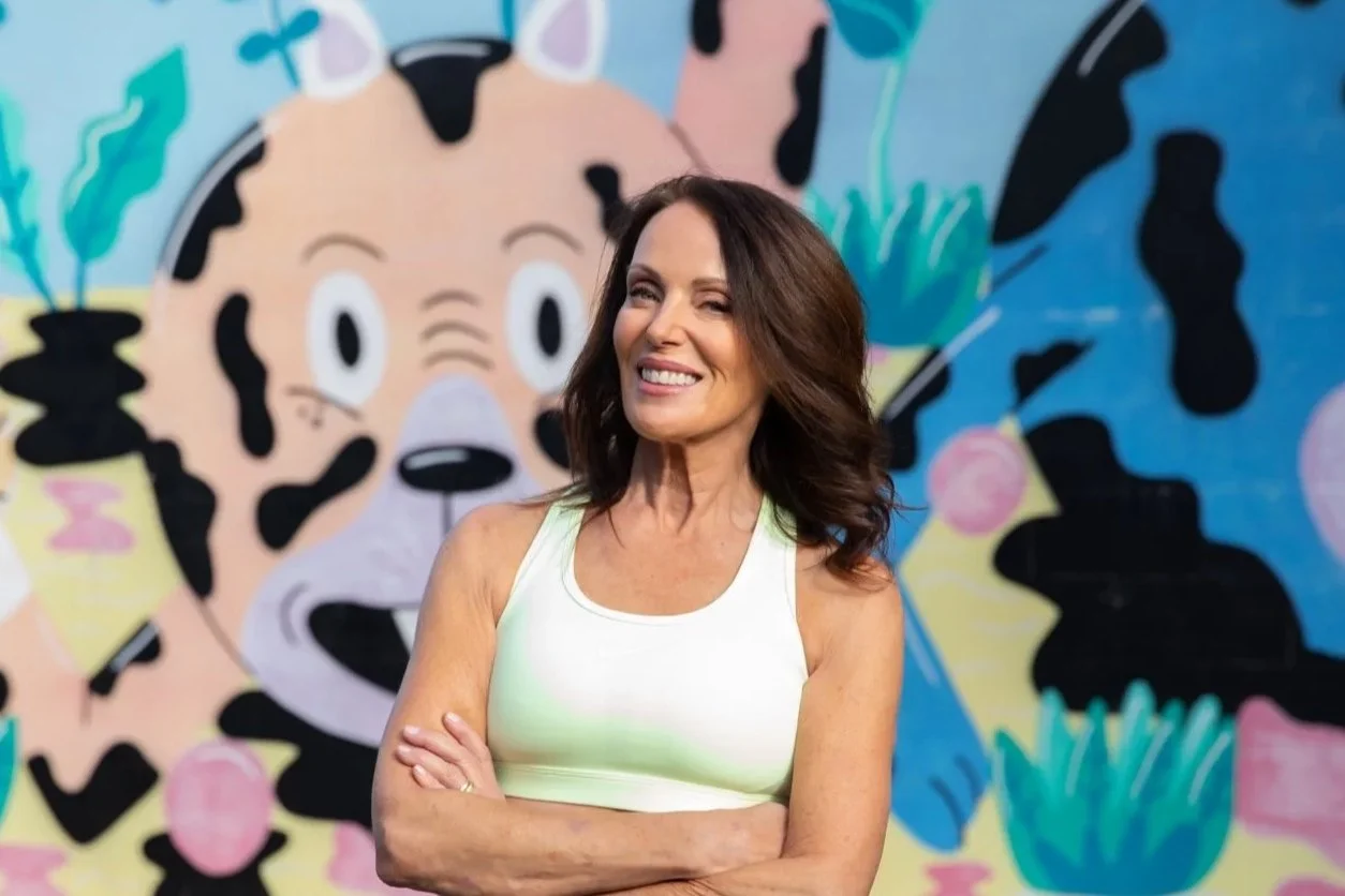 A smiling woman with dark brown hair in a white tank top standing in front of a colorful mural of a dog with big eyes, surrounded by abstract shapes and plants.