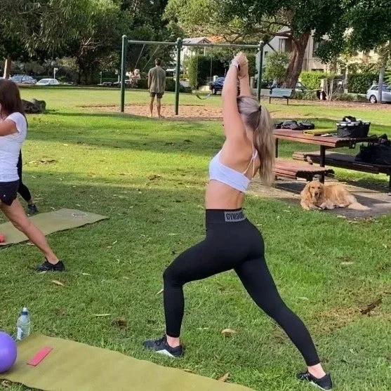 A woman with long hair in leggings and a sports bra practicing yoga in a park, with a dog lying on the ground nearby, and other people exercising in the background.