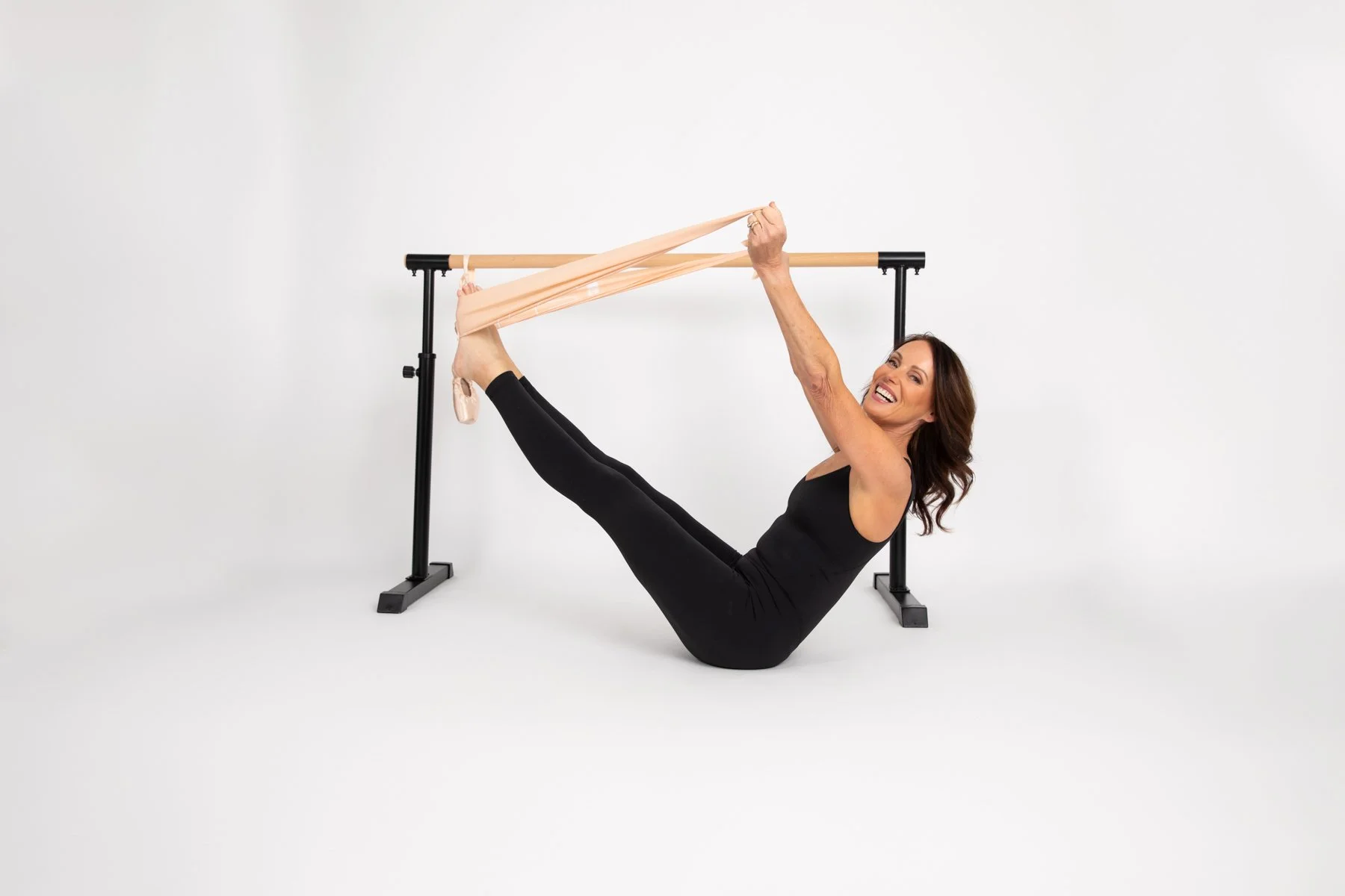 A smiling woman in black workout attire demonstrating a stretch with ballet tights while sitting on the floor in front of a ballet barre.