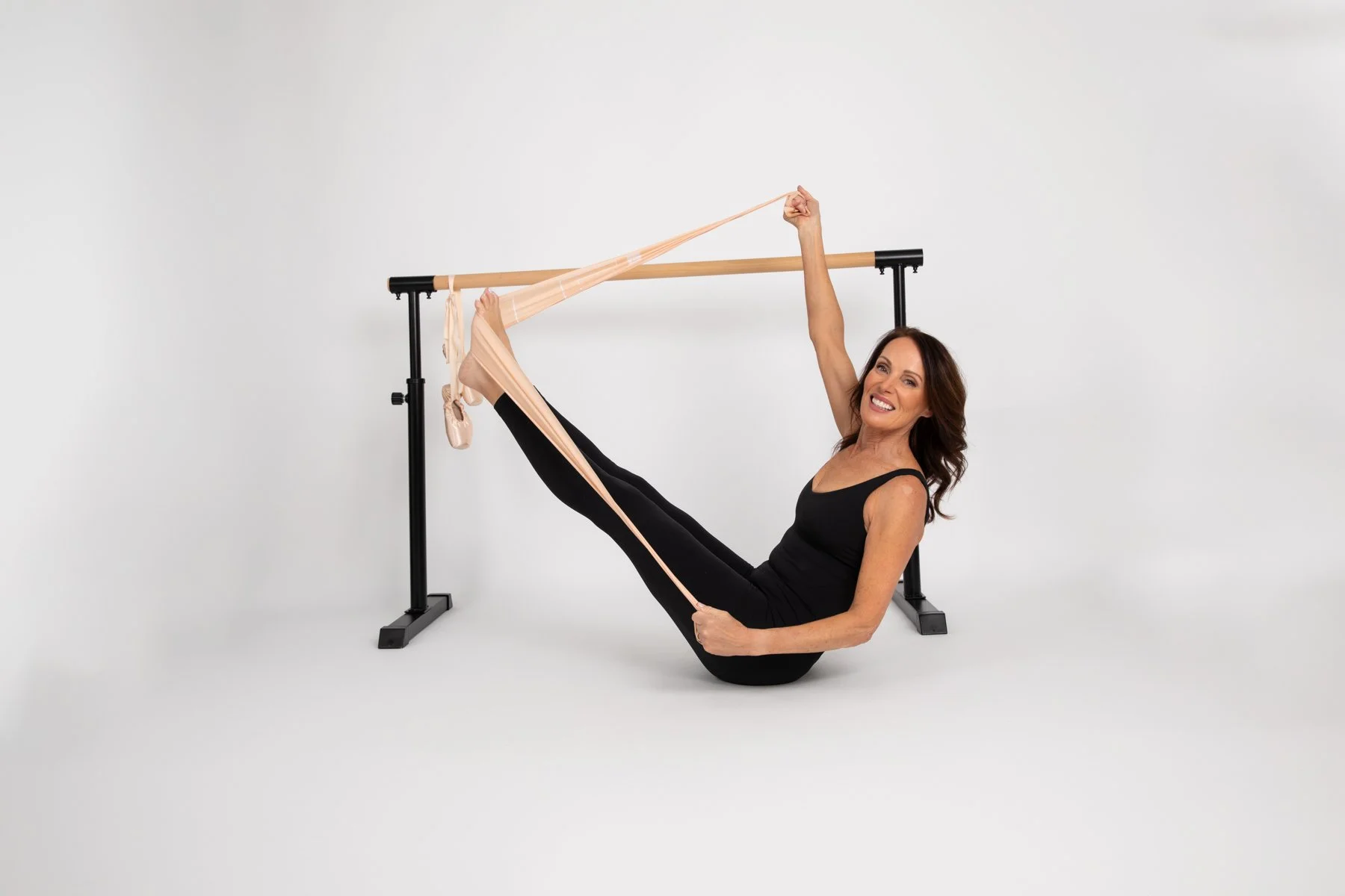 A woman in a black workout outfit practicing ballet poses with a resistance band, holding onto a ballet barre.