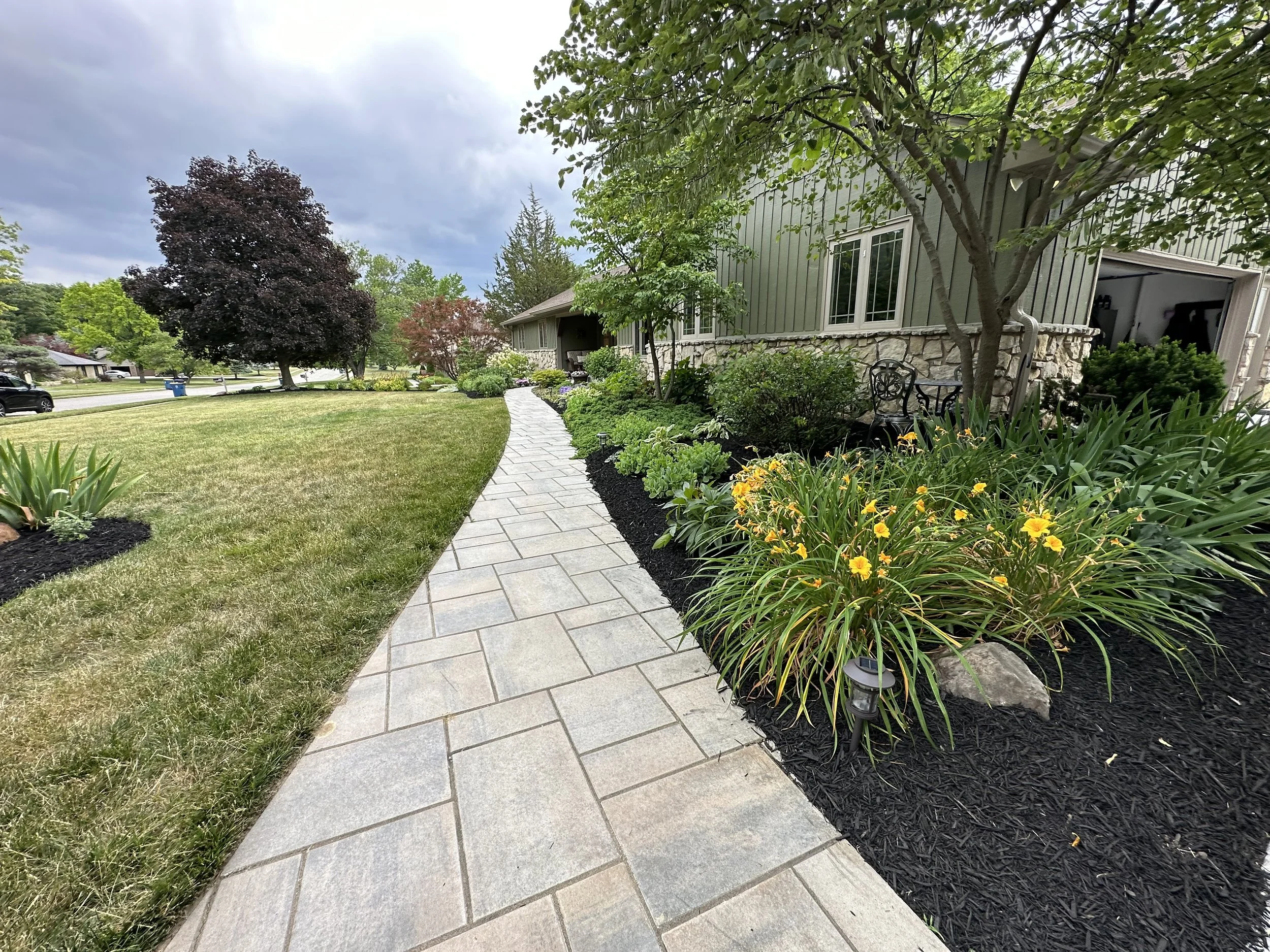 A curved stone pathway runs through a well-maintained front yard with green grass, flowering plants, trees, and a house with siding and large windows on the right side.