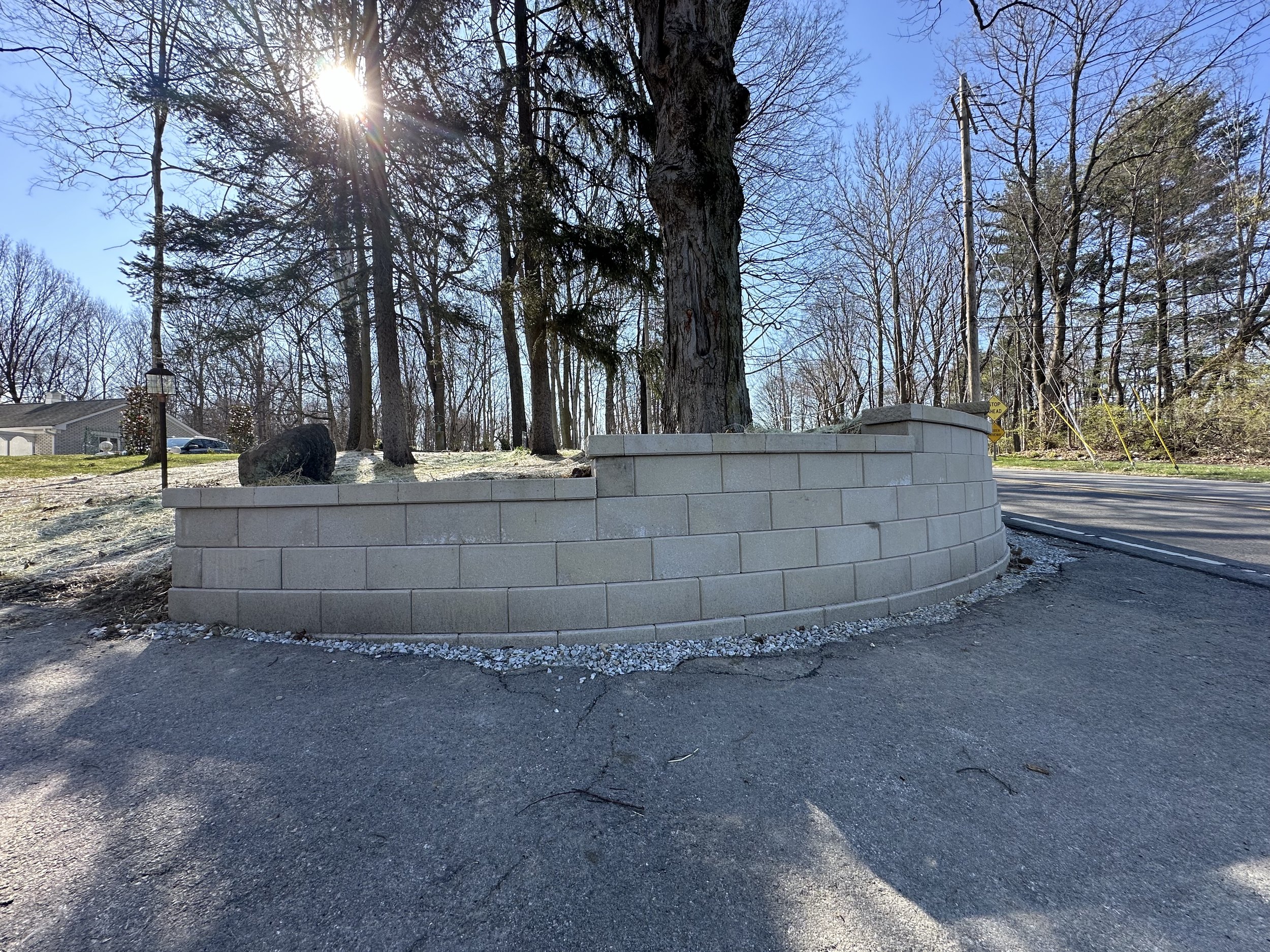 A landscaped roadside area with a new beige brick retaining wall surrounding a large tree, with sunlight shining through the branches, a paved road on the right, and a house with a car in the distance under a blue sky.