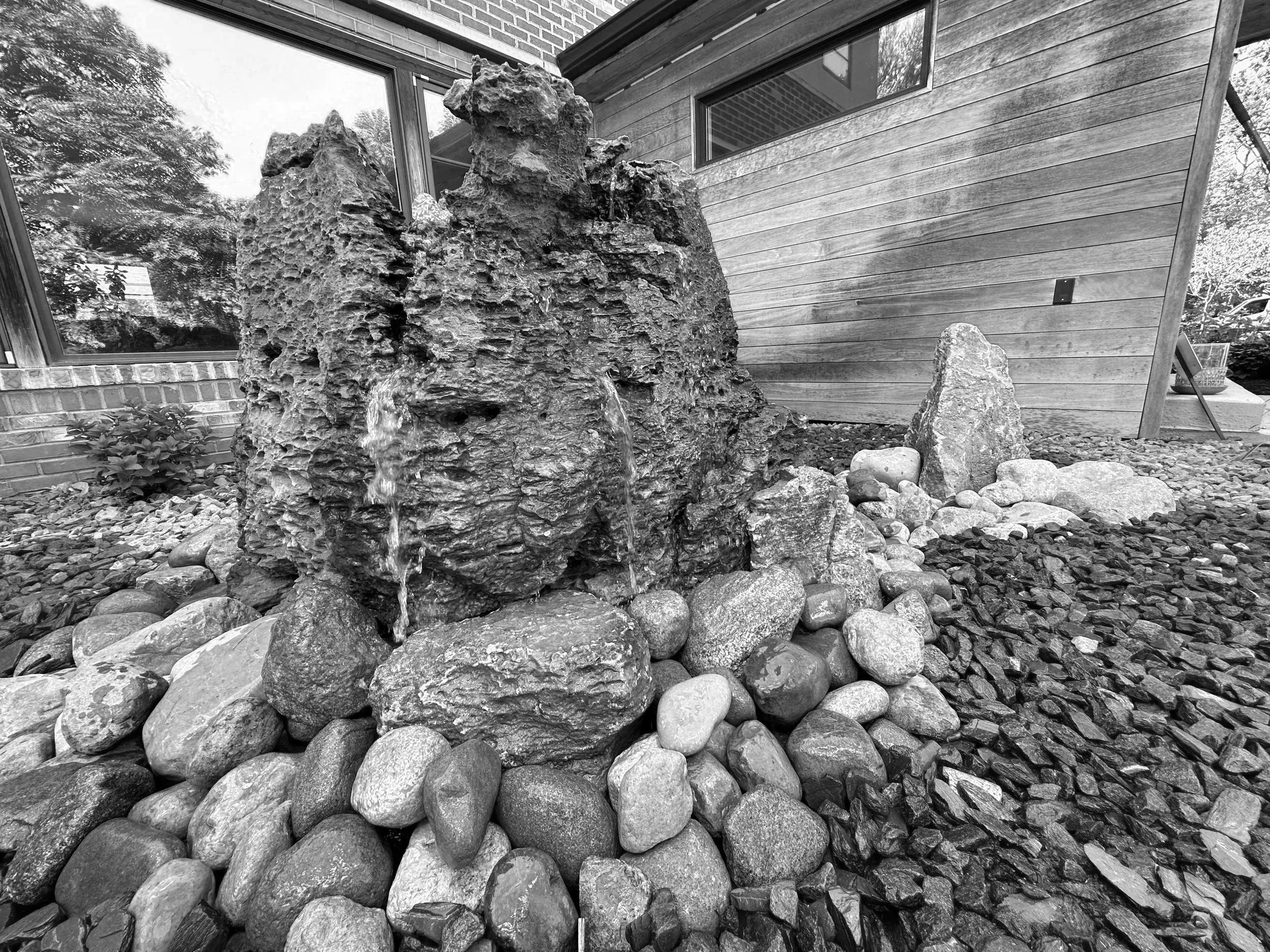 A black and white photo of a garden rock fountain with water flowing from small openings in a large, textured rock, surrounded by smaller rounded stones, positioned in front of a modern house with wood and brick exterior walls and large windows.