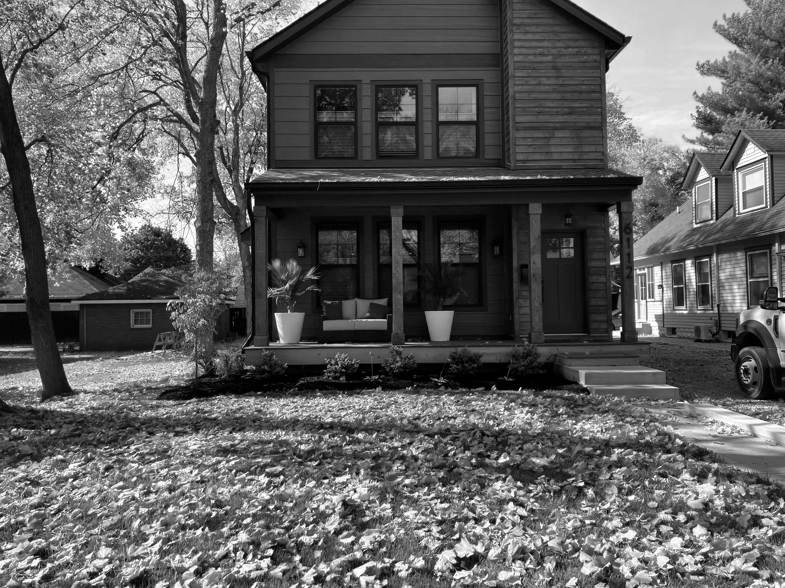 A black and white image of a two-story house with a front porch, two potted plants, and a sofa on the porch. There are trees with bare branches and fallen leaves on the ground, indicating autumn. The house number is visible on a column.