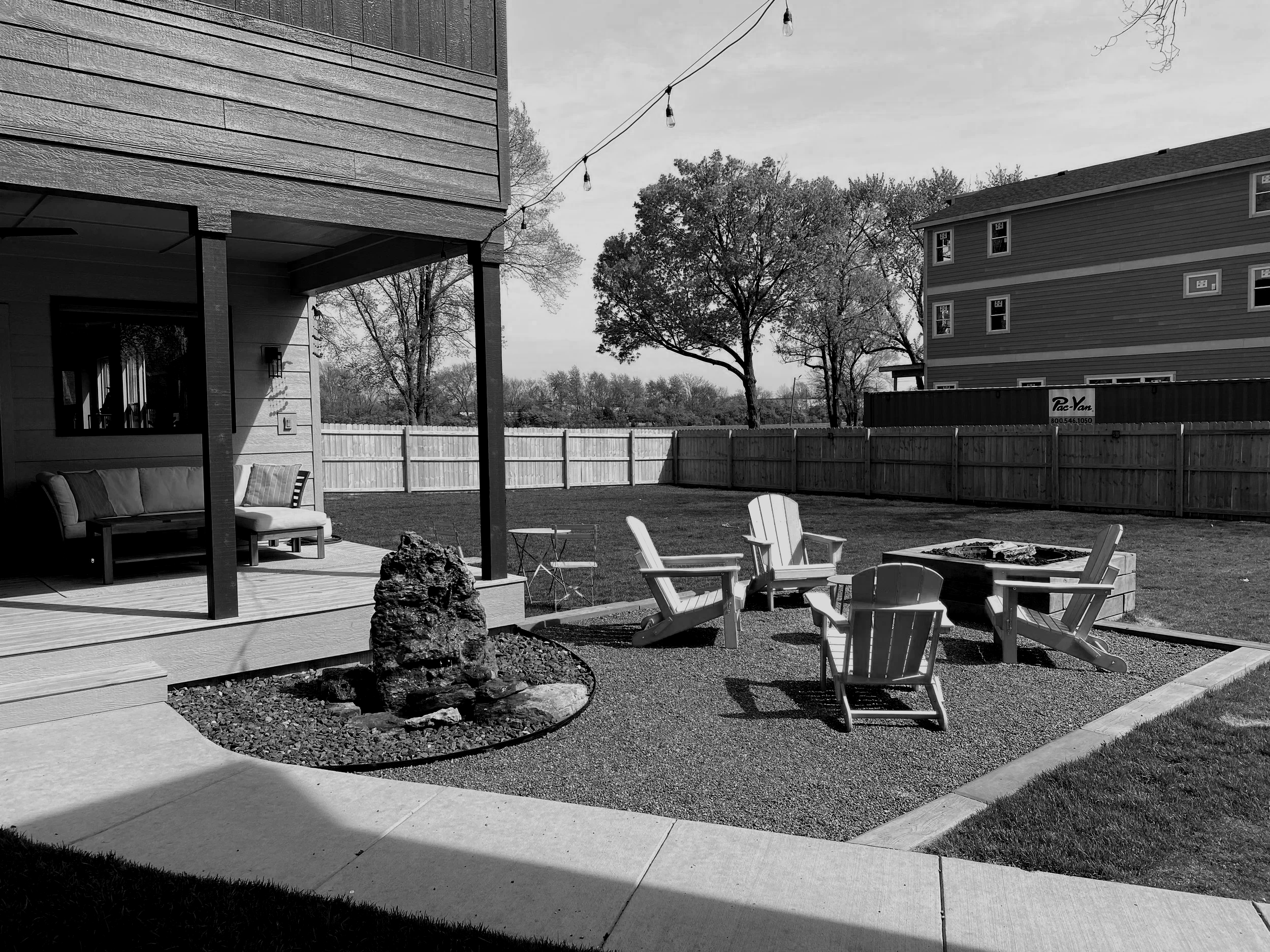 A backyard patio with Adirondack chairs around a fire pit, a house with a porch, string lights, and a fenced yard with trees in the background.