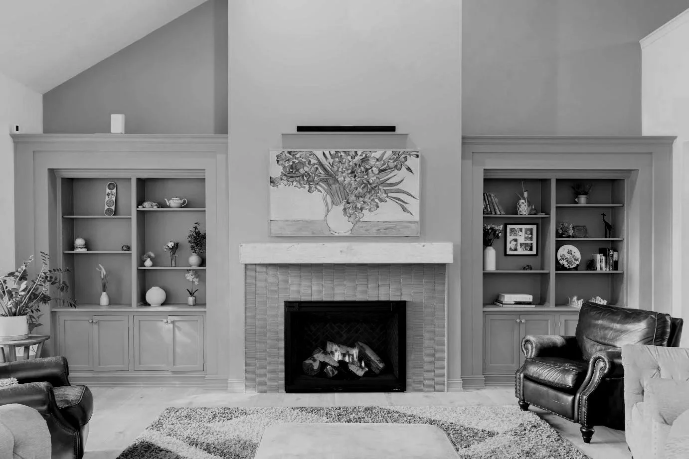 Living room with built-in shelves on either side of a fireplace, decorated with vases, books, and ornaments; a large painting of flowers above the fireplace; black leather armchairs; a window to the left with plants; a rug on the floor.