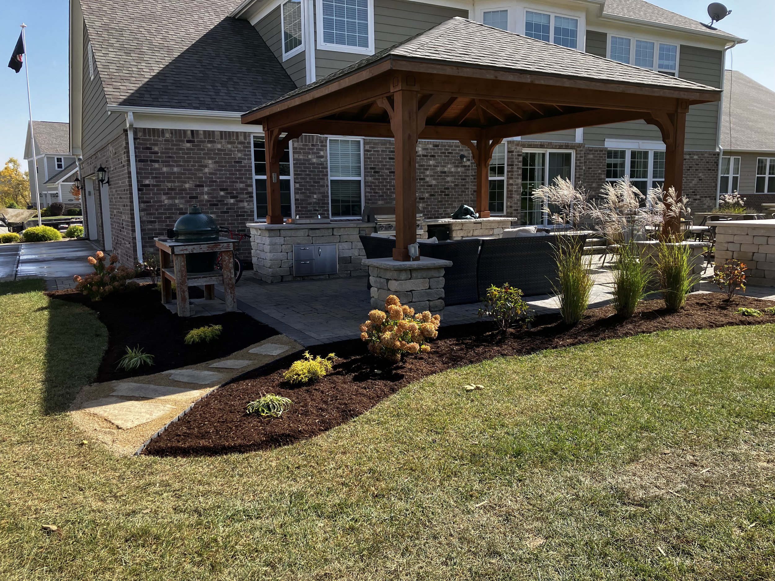 A backyard patio with a wooden pergola, outdoor seating, a stone firepit, a grill, and landscaped flower beds with plants and mulch.