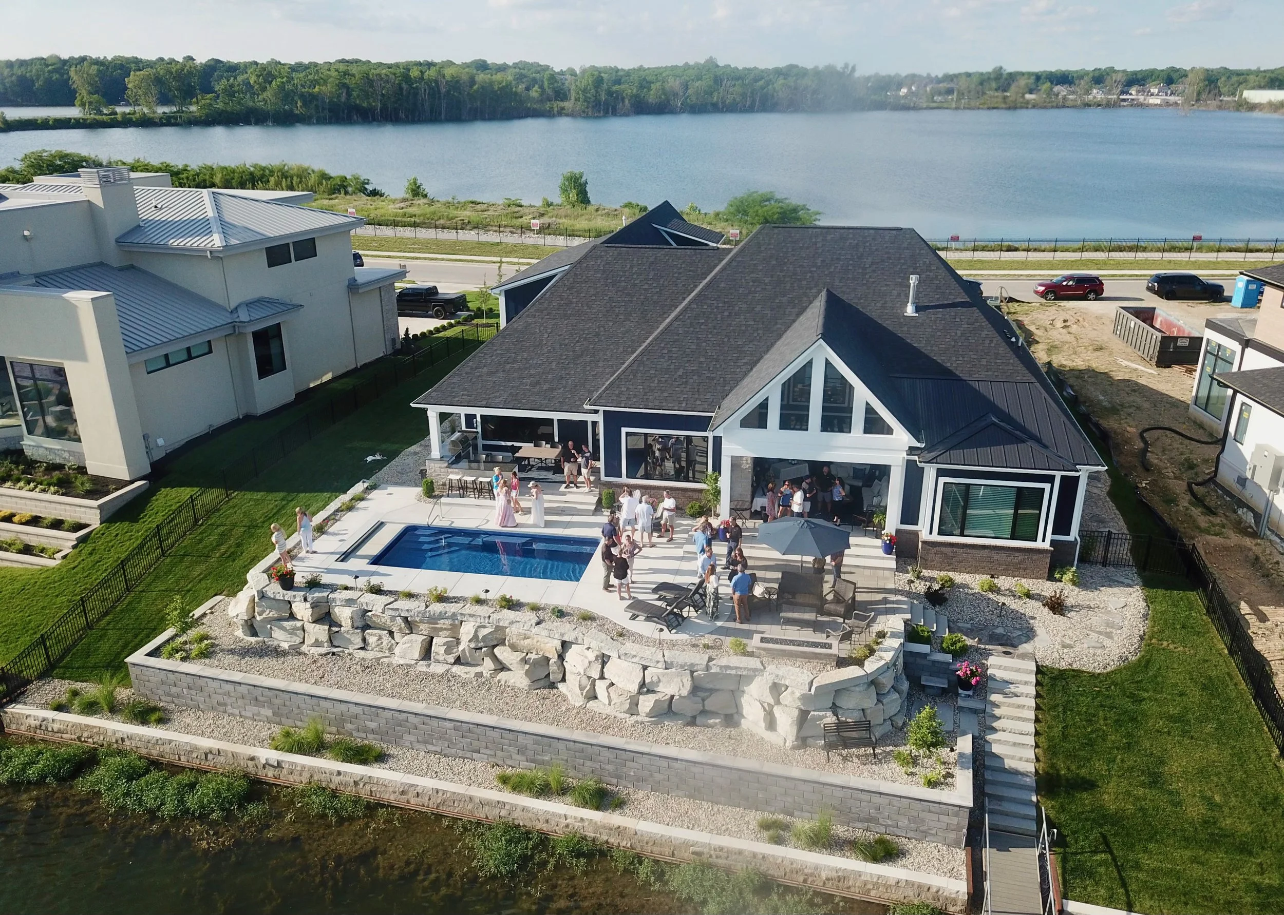 Aerial view of a modern house with a backyard pool, shaded patio, and a gathering of people, overlooking a large lake.