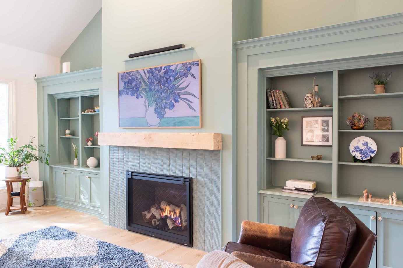 Living room with a light blue wall, built-in bookshelves, a fireplace with a wooden mantel, a blue floral painting above the fireplace, a leather armchair, a patterned rug, and a potted plant near the window.