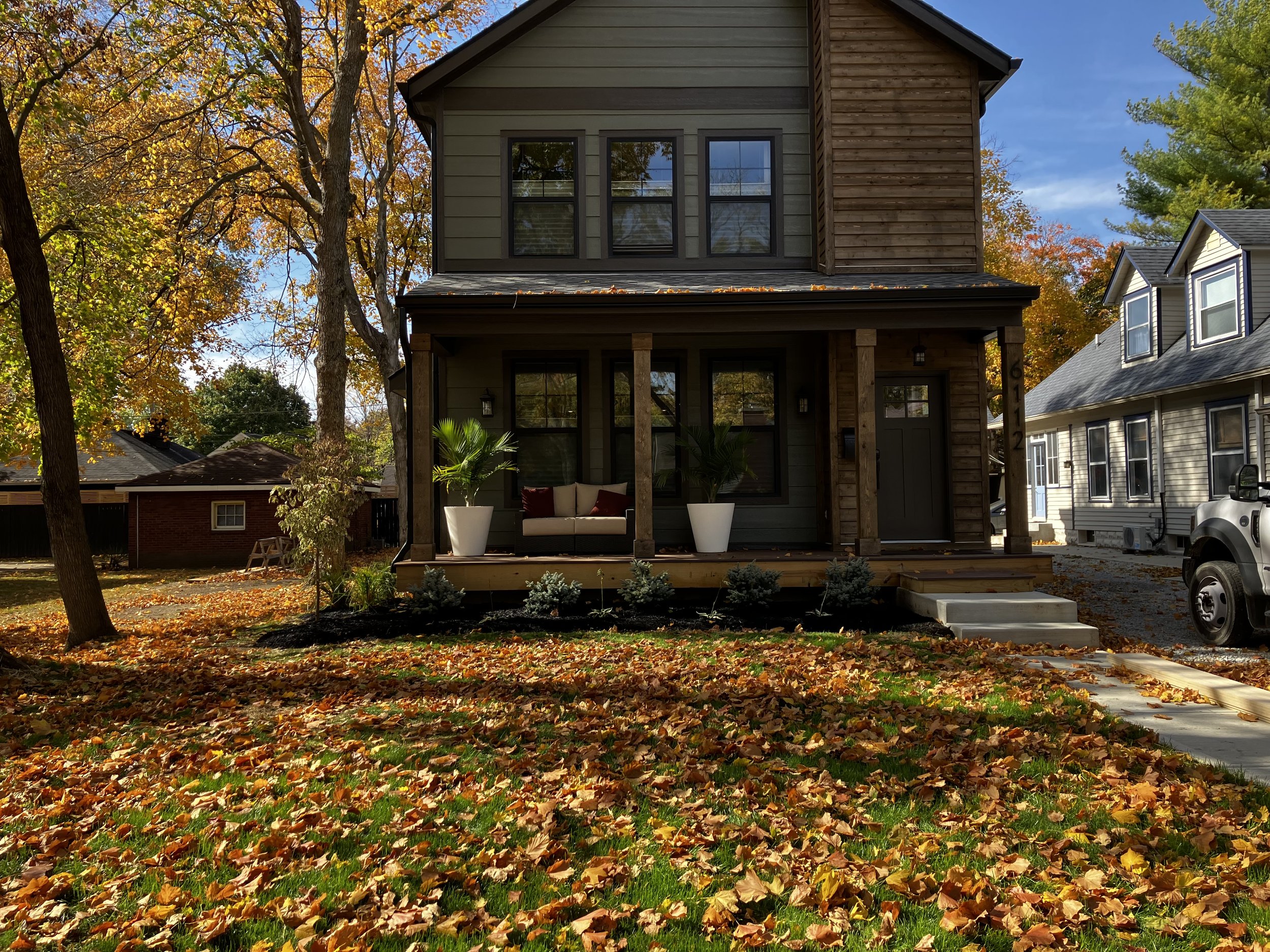 A two-story house with a front porch, during autumn with fallen leaves on the ground, trees with orange and green leaves, and neighboring houses visible.