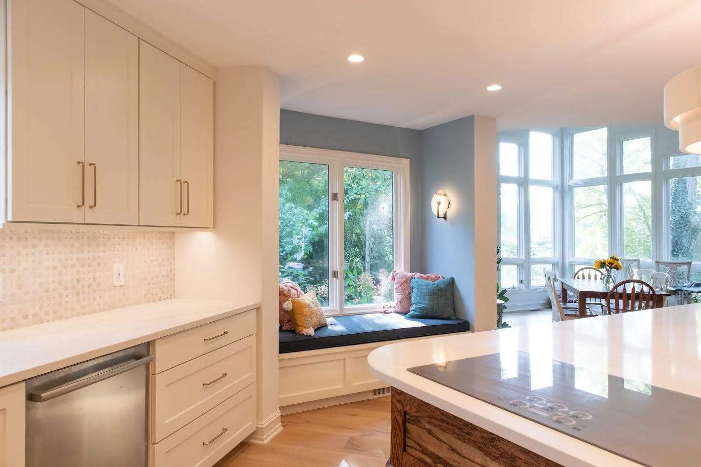 Kitchen with white cabinets, a window seat with cushions, a dining table with chairs, and large bay windows with a view of trees outside.
