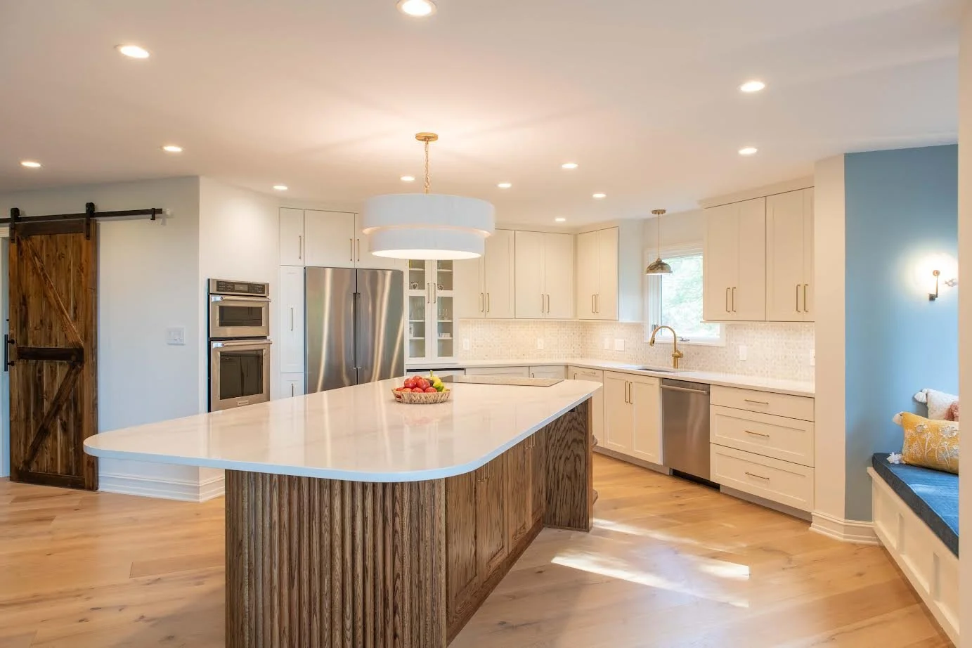 Modern kitchen with white cabinets, stainless steel appliances, a large island with a wooden base and white countertop, and a window with a built-in bench seat and yellow cushion.