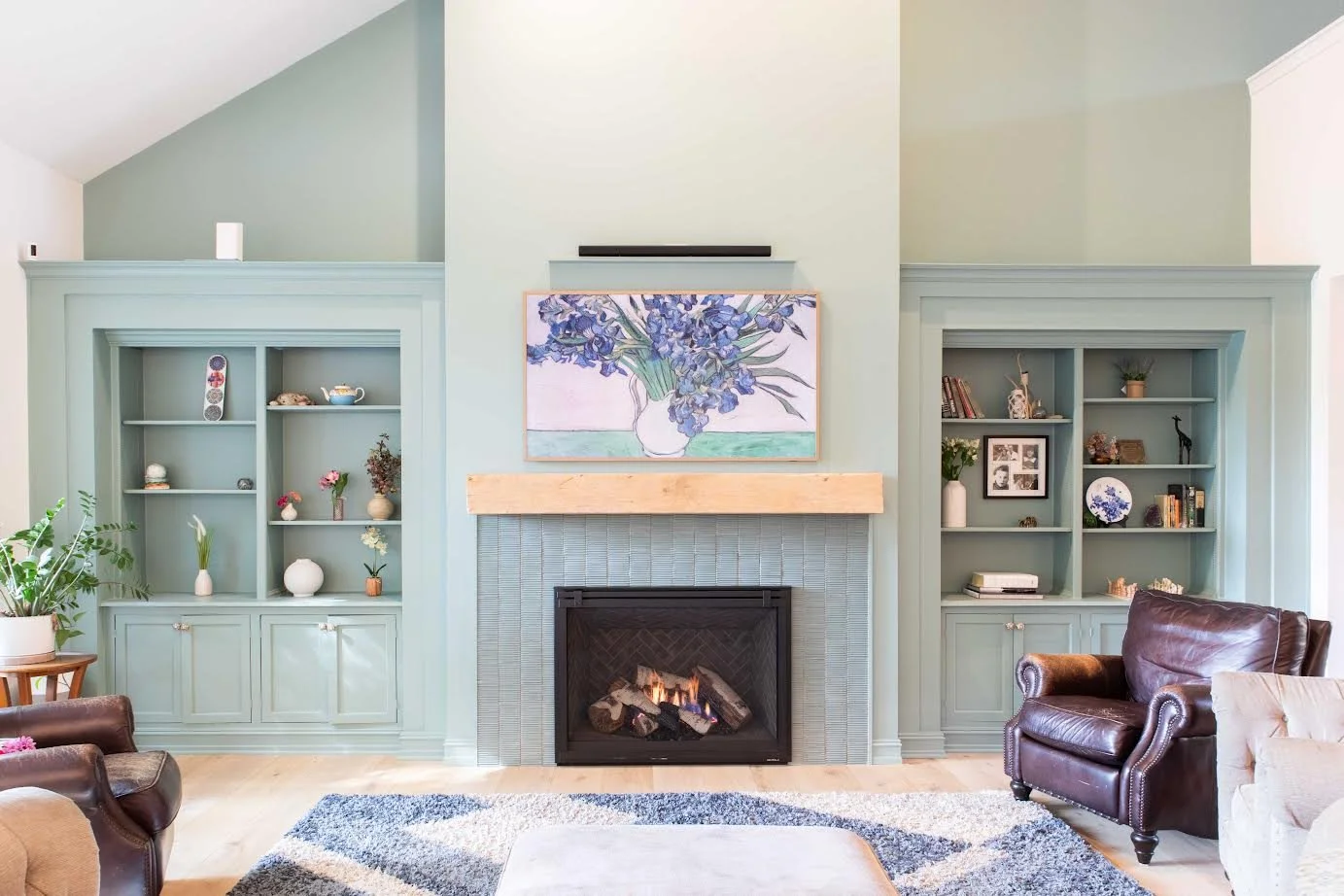 Living room with a fireplace, pastel green walls, built-in bookshelves with decorative items, a marble fireplace mantel, a framed floral painting above the fireplace, brown leather chairs, a multicolored rug, and potted plants.