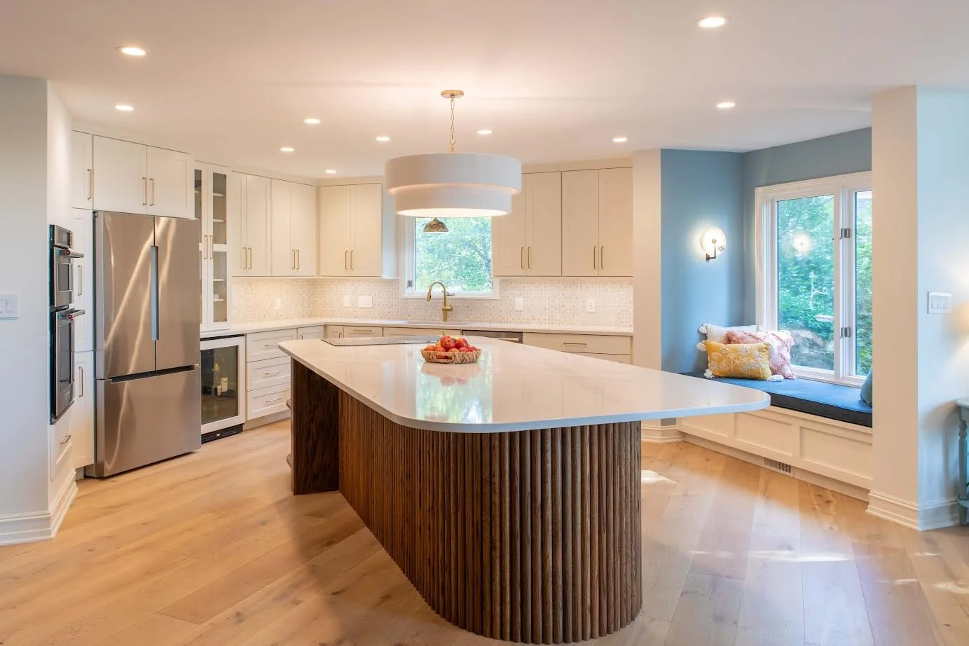 Modern kitchen with white cabinets, a large central island with a curved wooden base and white countertop, stainless steel appliances, a window with a seating nook, and decorative pillows.