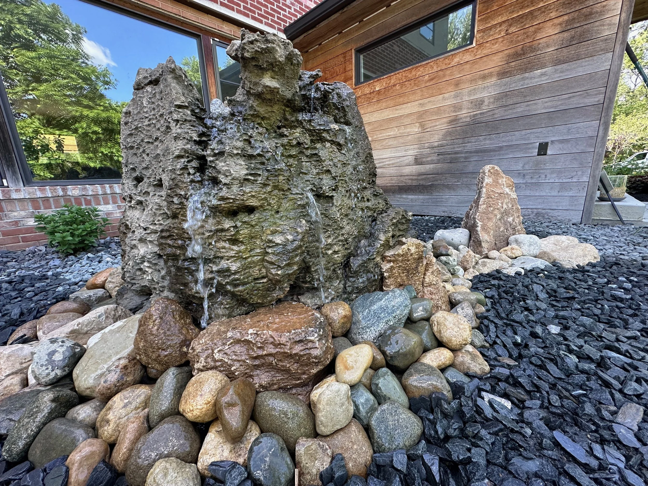 A landscaped garden with a rock water fountain surrounded by smooth river stones, with a house featuring brick and wooden exterior walls in the background.