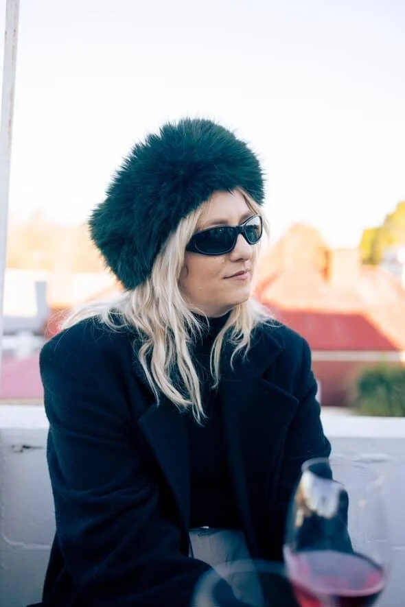 A woman with blonde hair wearing black sunglasses, a black fur hat, and a dark coat, sitting outdoors with a blurred background of rooftops and sky.