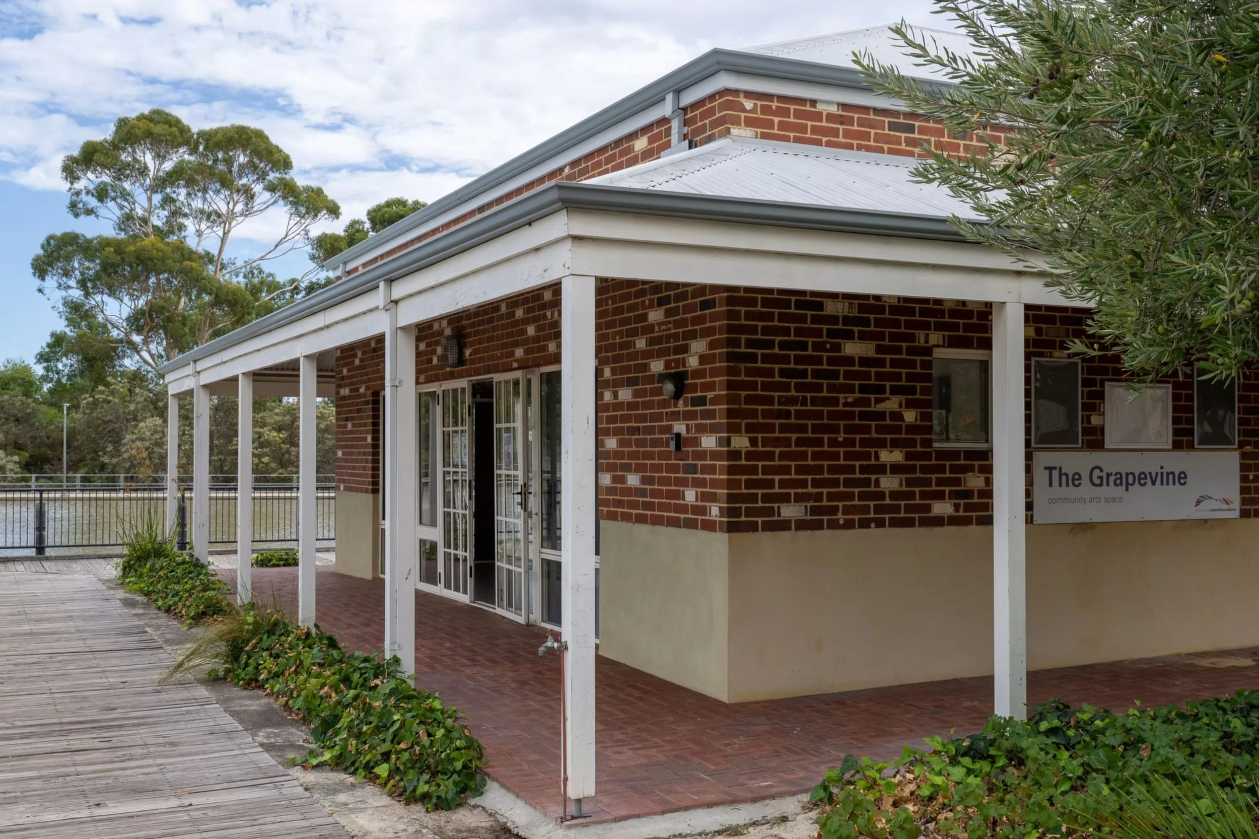 ID: An outside corner of The Grapevine. It has red bricks and a dark coloured roof. Bushes line the edges.